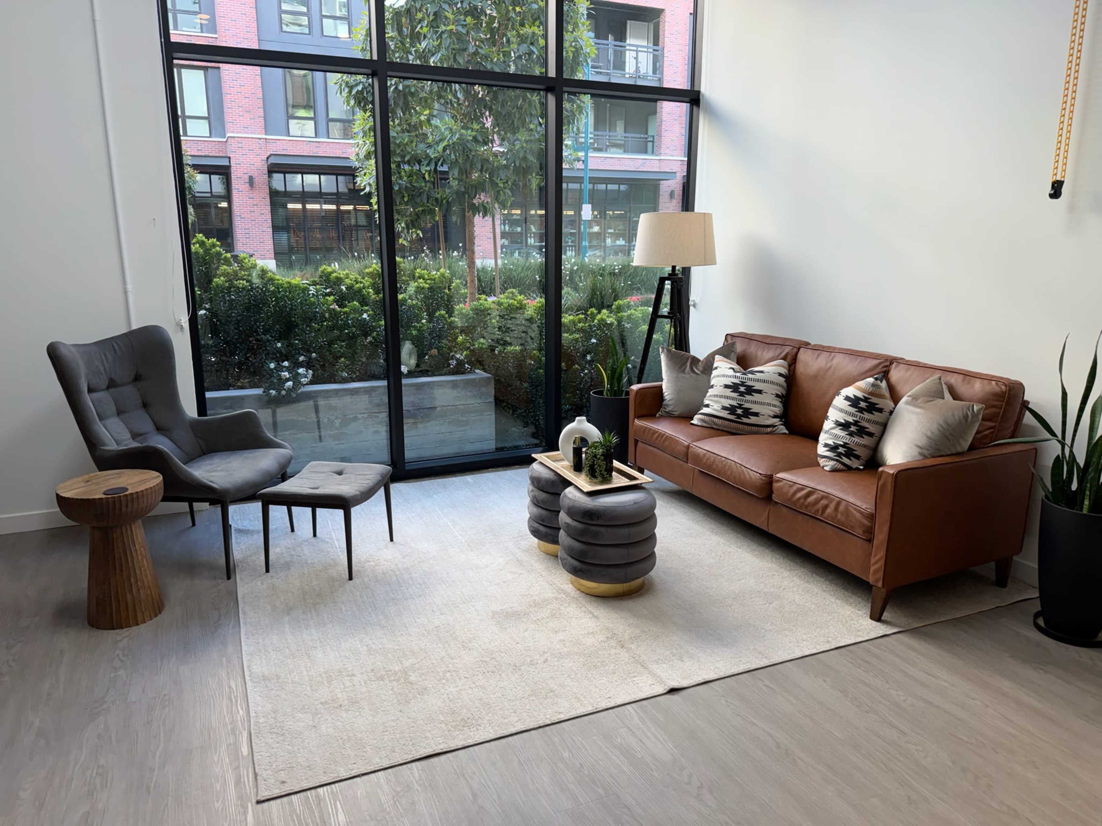 The scene features a modern living room with a brown leather sofa, a gray armchair, a wooden side table, and a coffee table on a light area rug, set against a large window overlooking greenery.