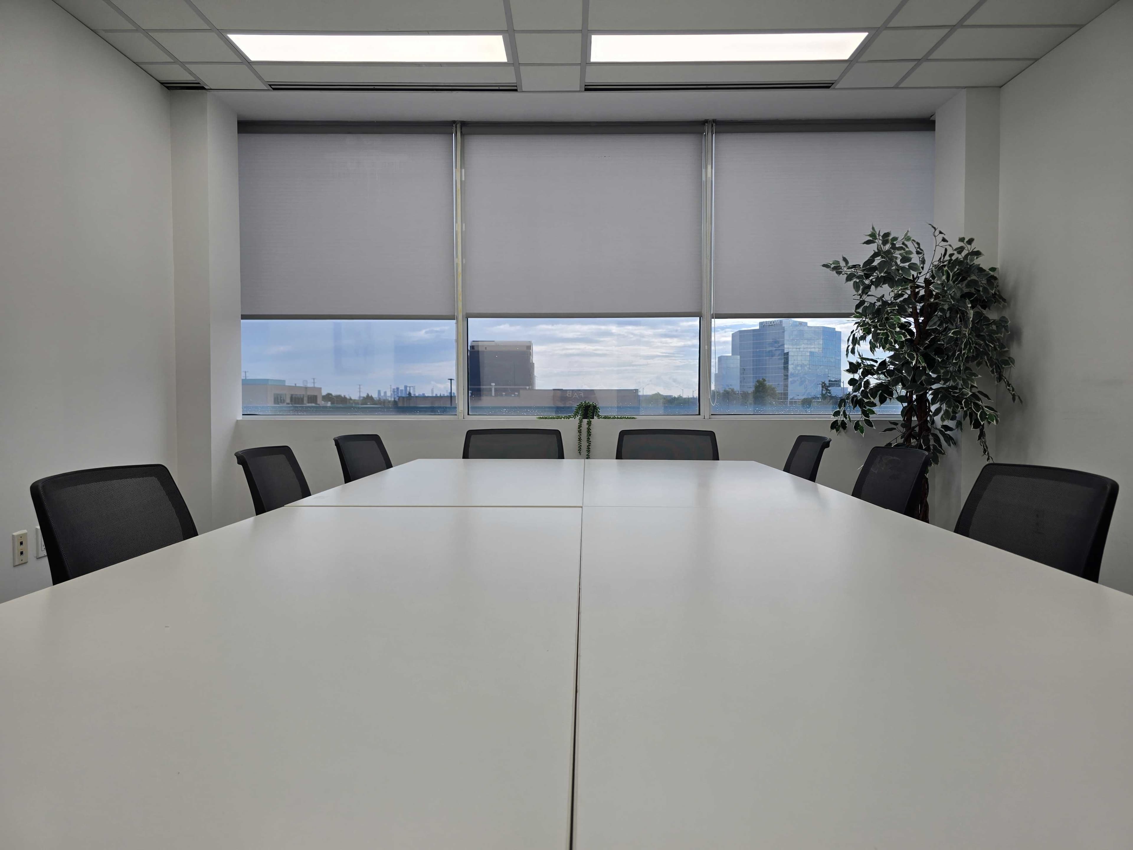 A conference room features a long table surrounded by black chairs, with large windows showing an outdoor view.