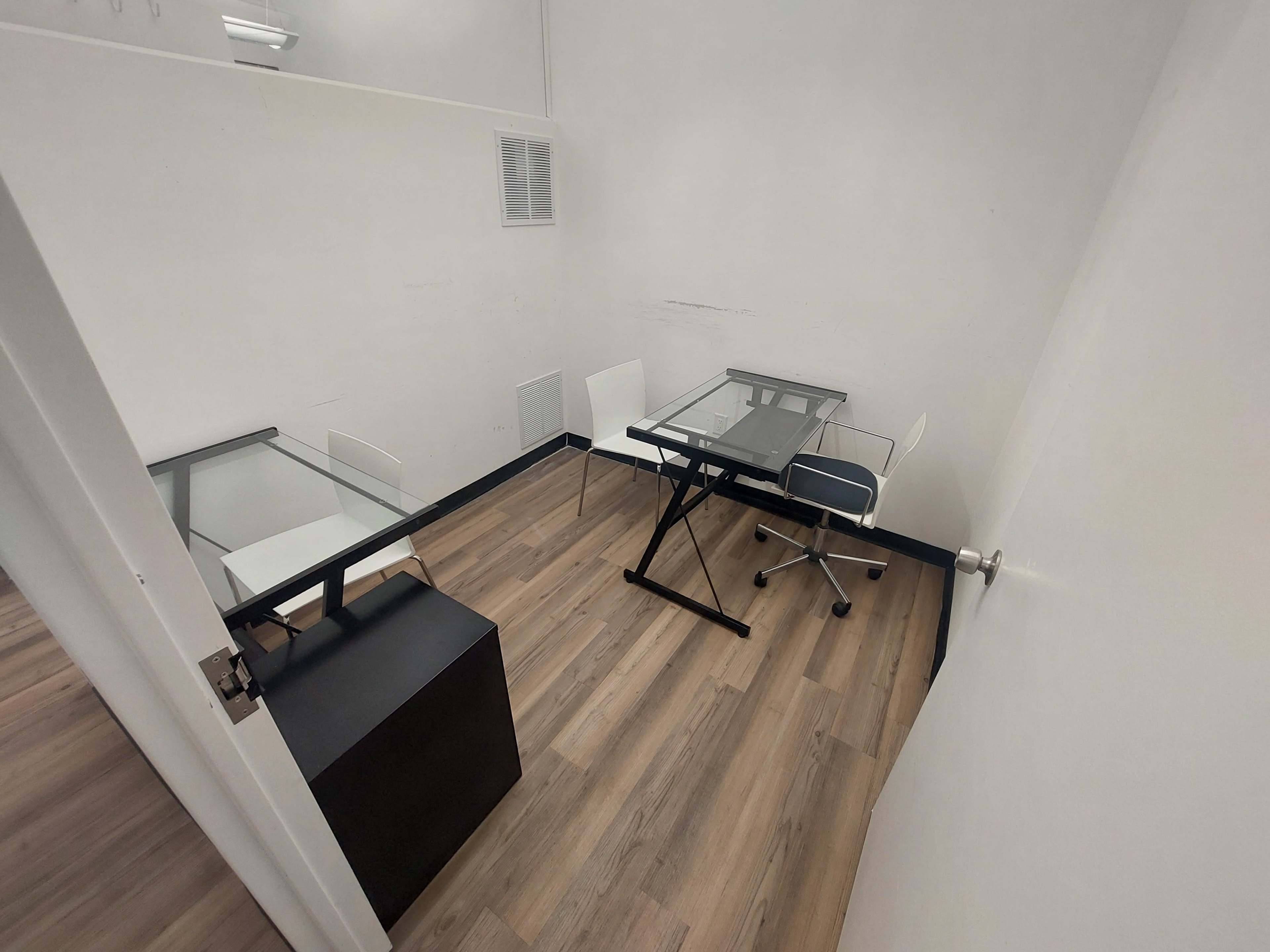 The image shows a small office space with two glass-top desks and three chairs arranged on a wooden floor.