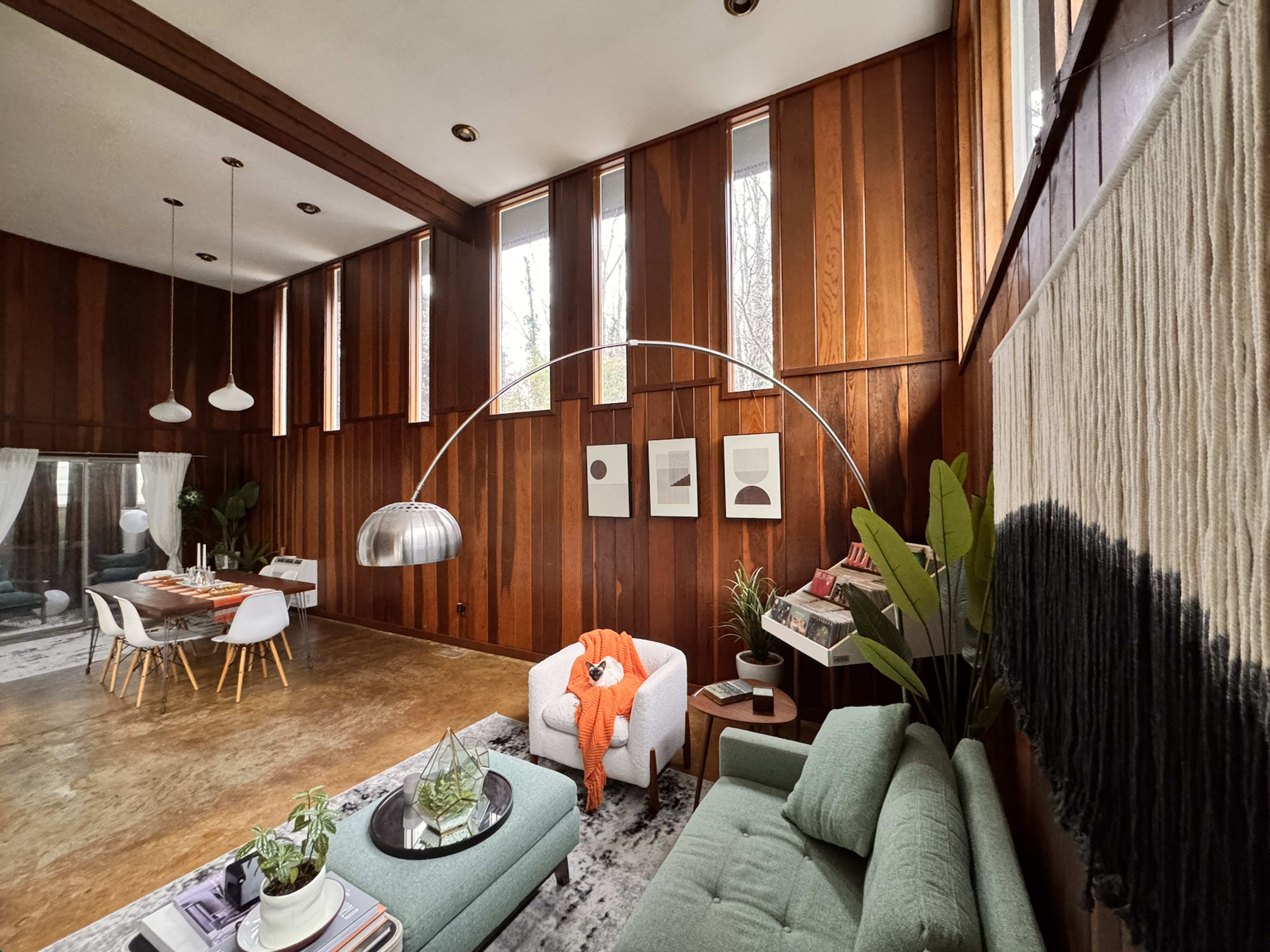 The image shows a modern living room with wood-paneled walls, large windows, a green sofa, a curved floor lamp, and a dining area in the background.