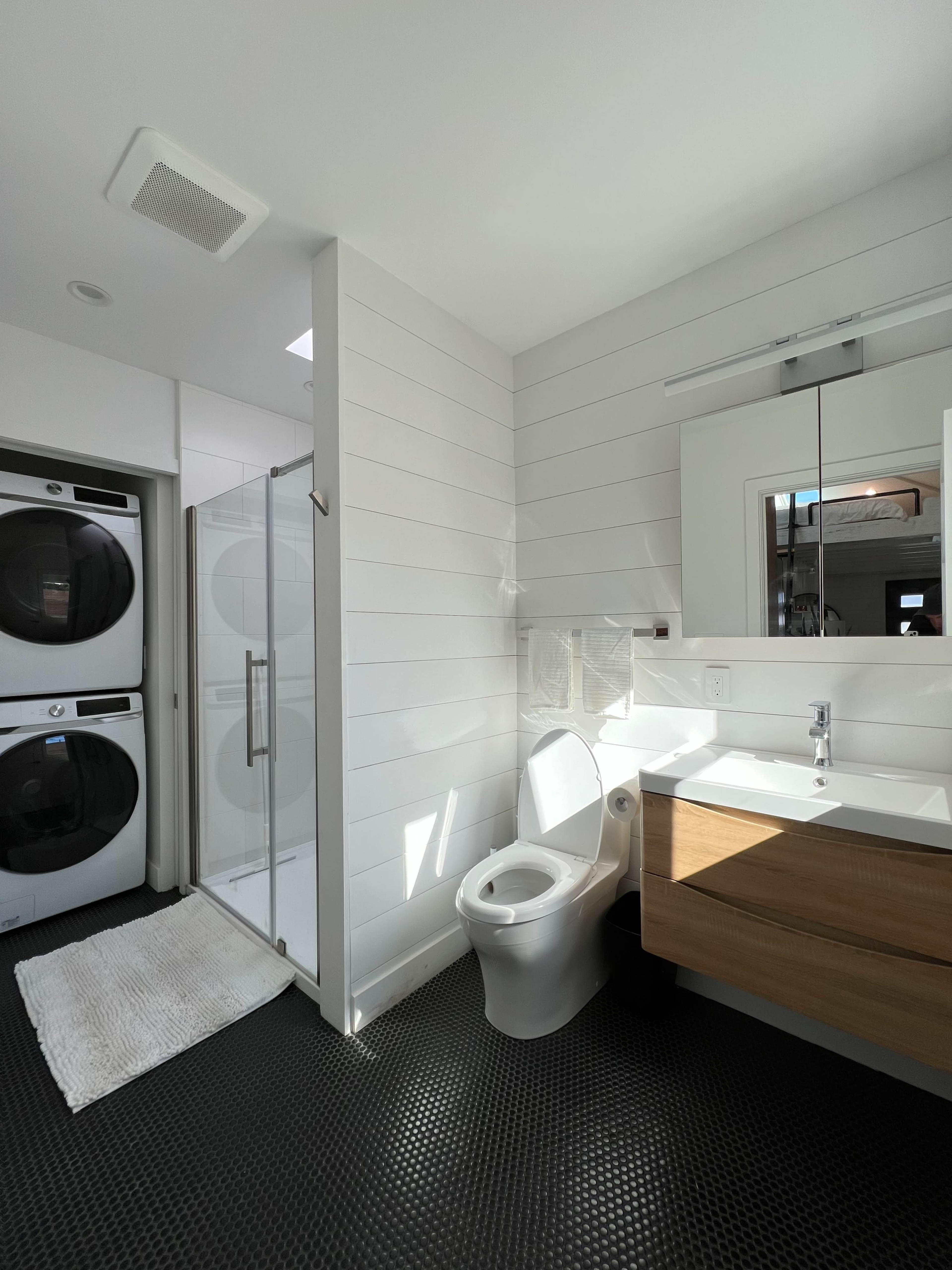 The image shows a modern bathroom featuring a toilet, a glass shower, a mirror above a wooden vanity, and a stacked washer and dryer.