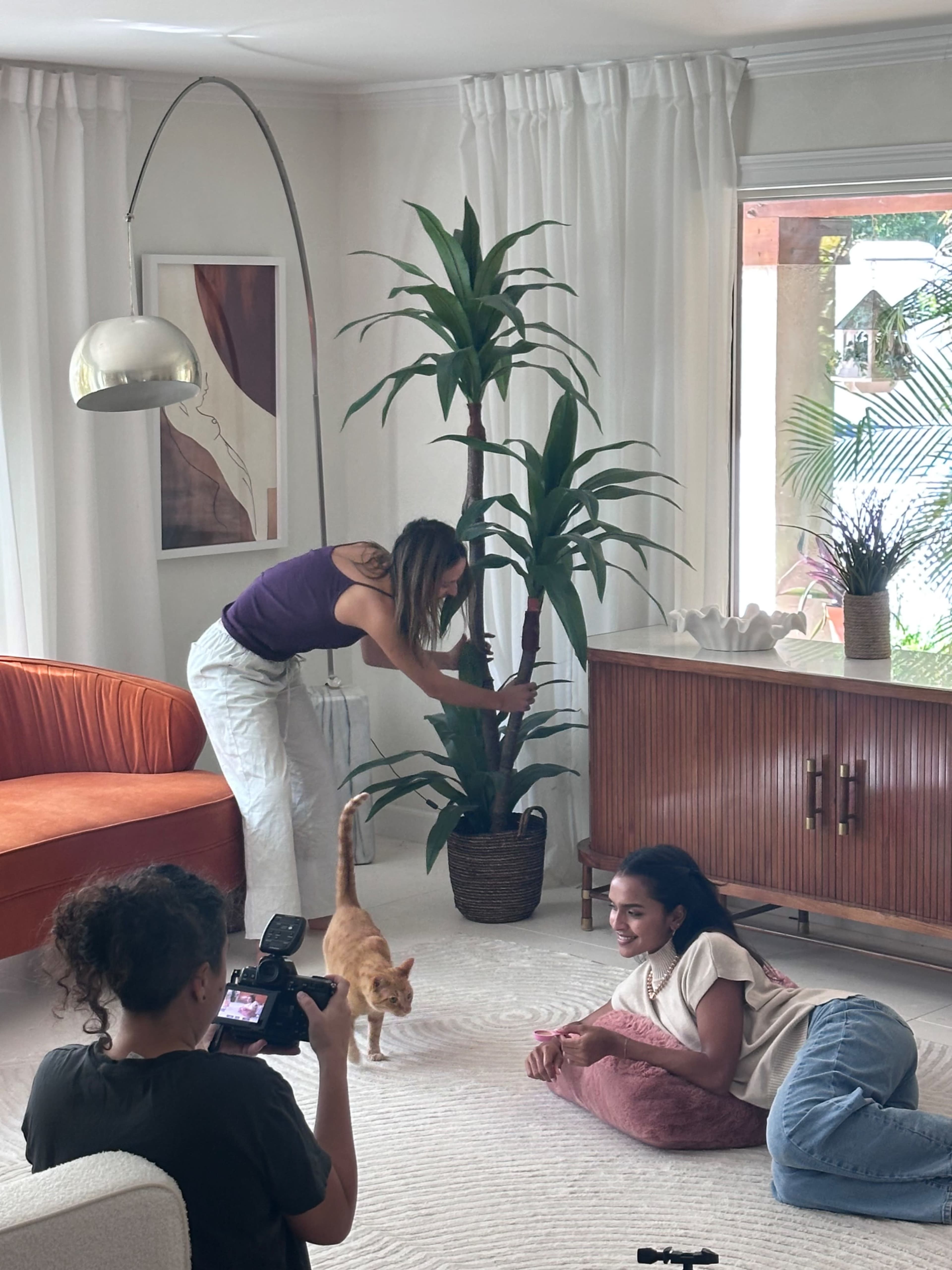 A woman adjusts a plant while another woman lies on the floor and a person with a camera captures the scene in a well-lit living room.