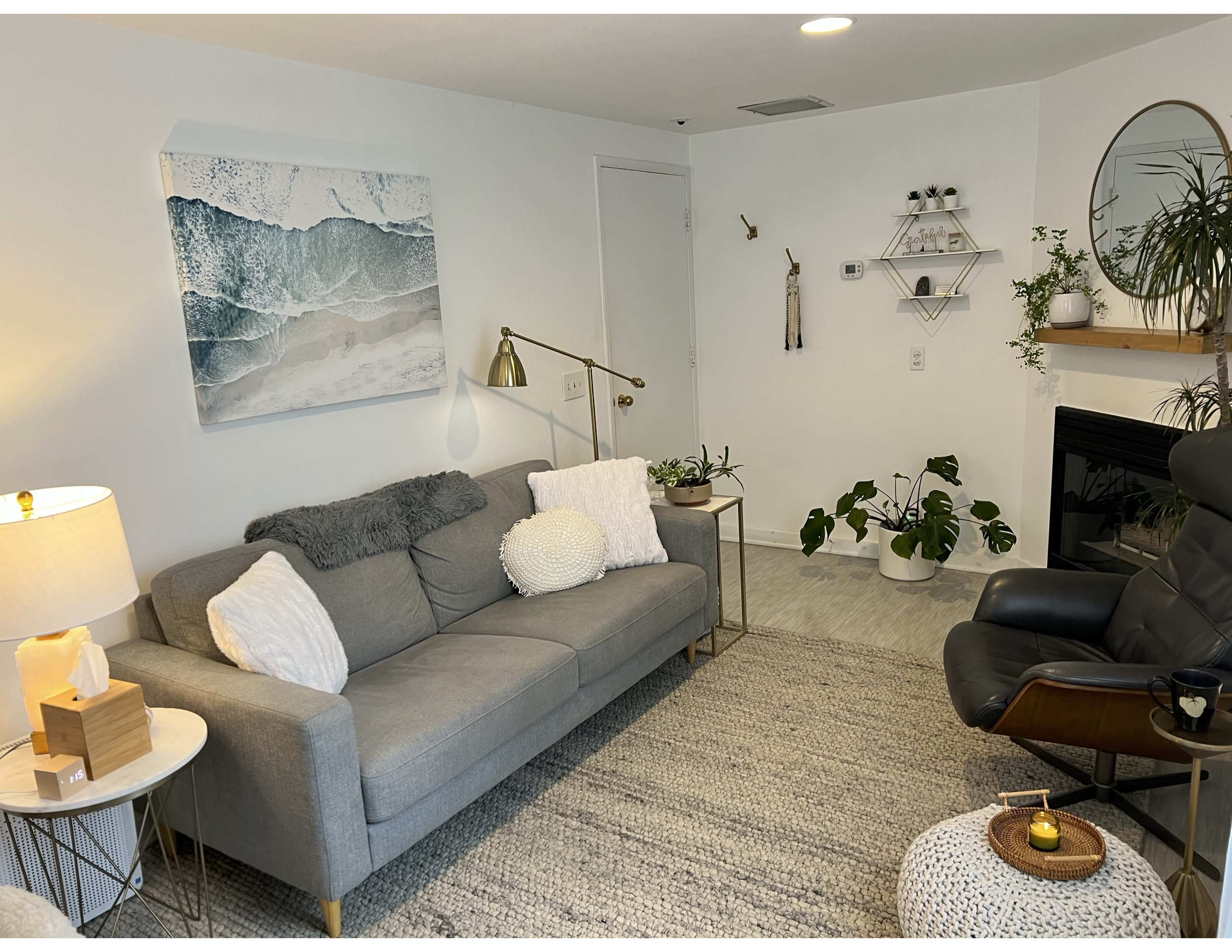 The image shows a cozy living room featuring a gray couch with decorative pillows, a floor lamp, a small round side table, and a black armchair, all complemented by a wall with a coastal-themed artwork and a plant shelf.