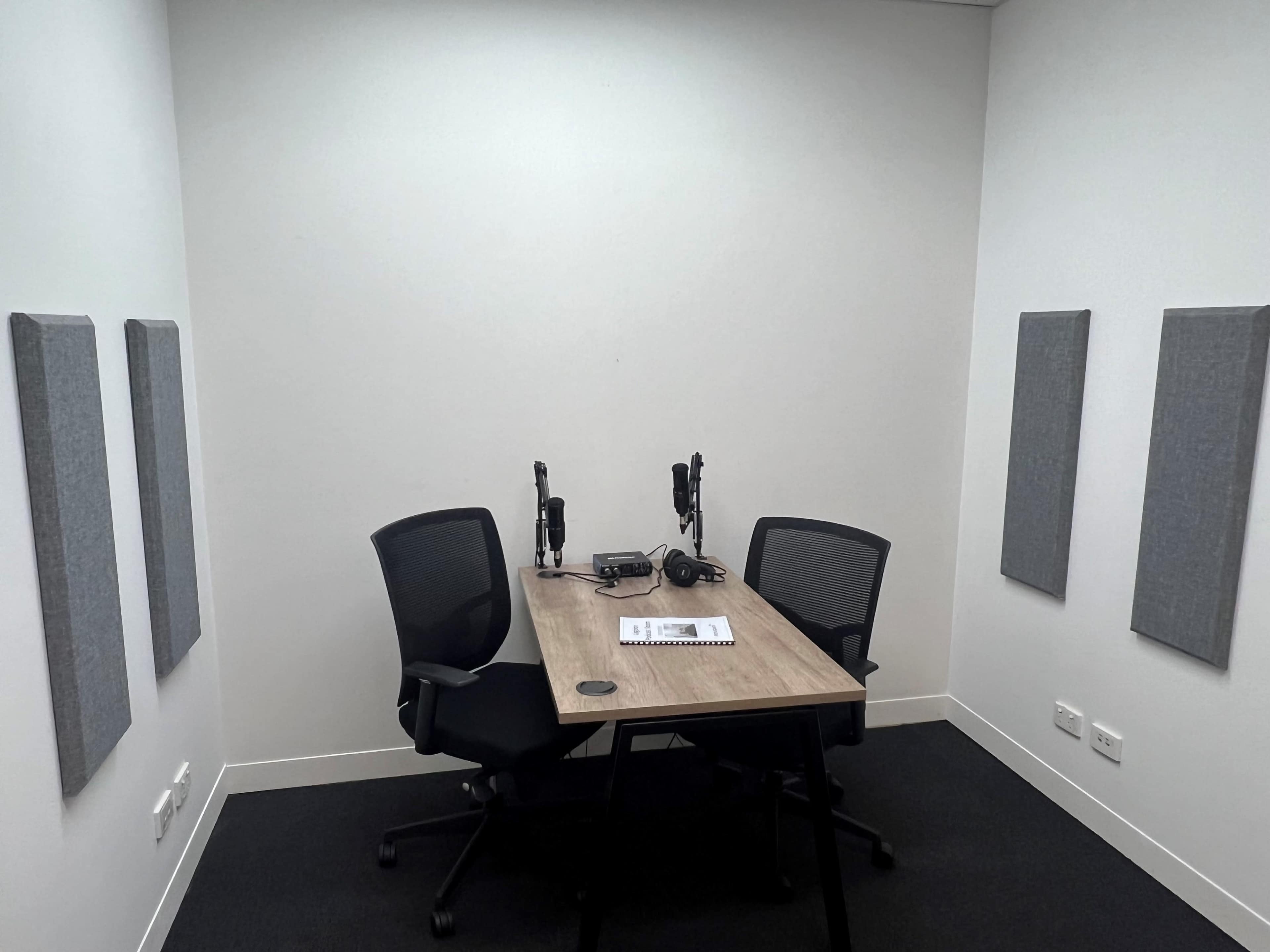 The image shows a small recording studio with a wooden table, two chairs, and soundproof panels on the walls.