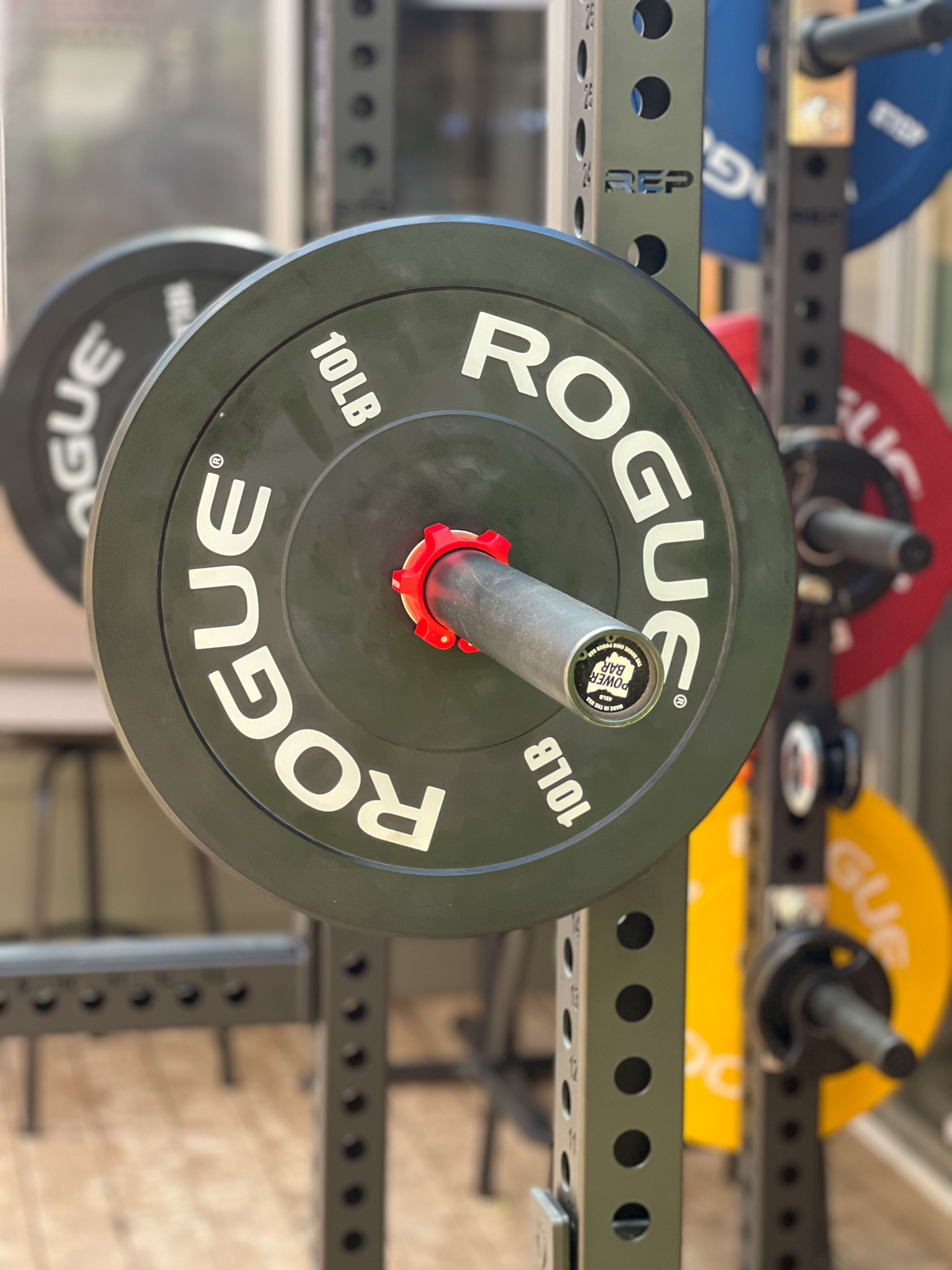 A barbell with a 10-pound Rogue weight plate is mounted on a weight rack in a gym.