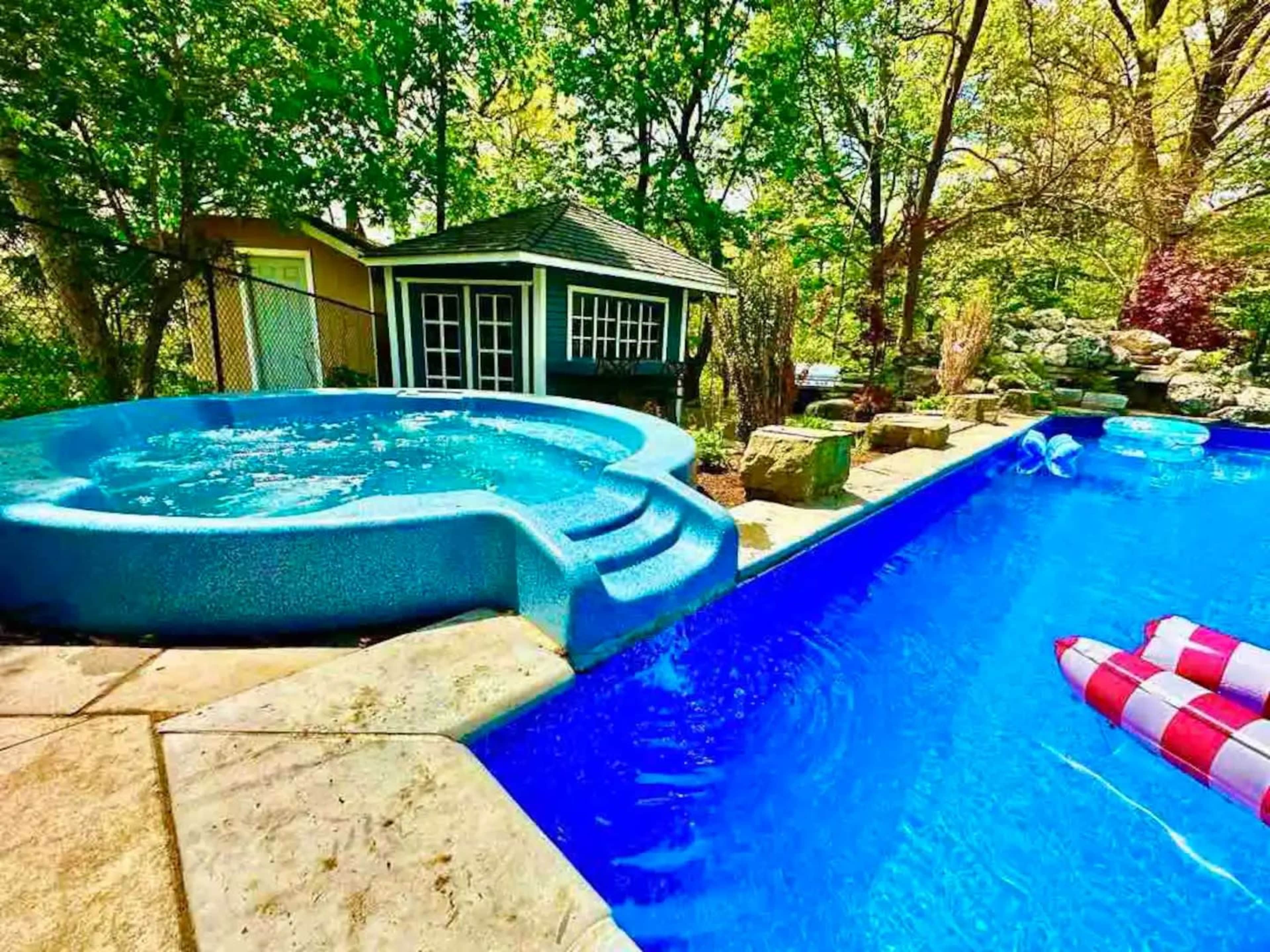 A backyard featuring a blue pool with a hot tub steps leading to it, surrounded by trees and a small shed in the background.