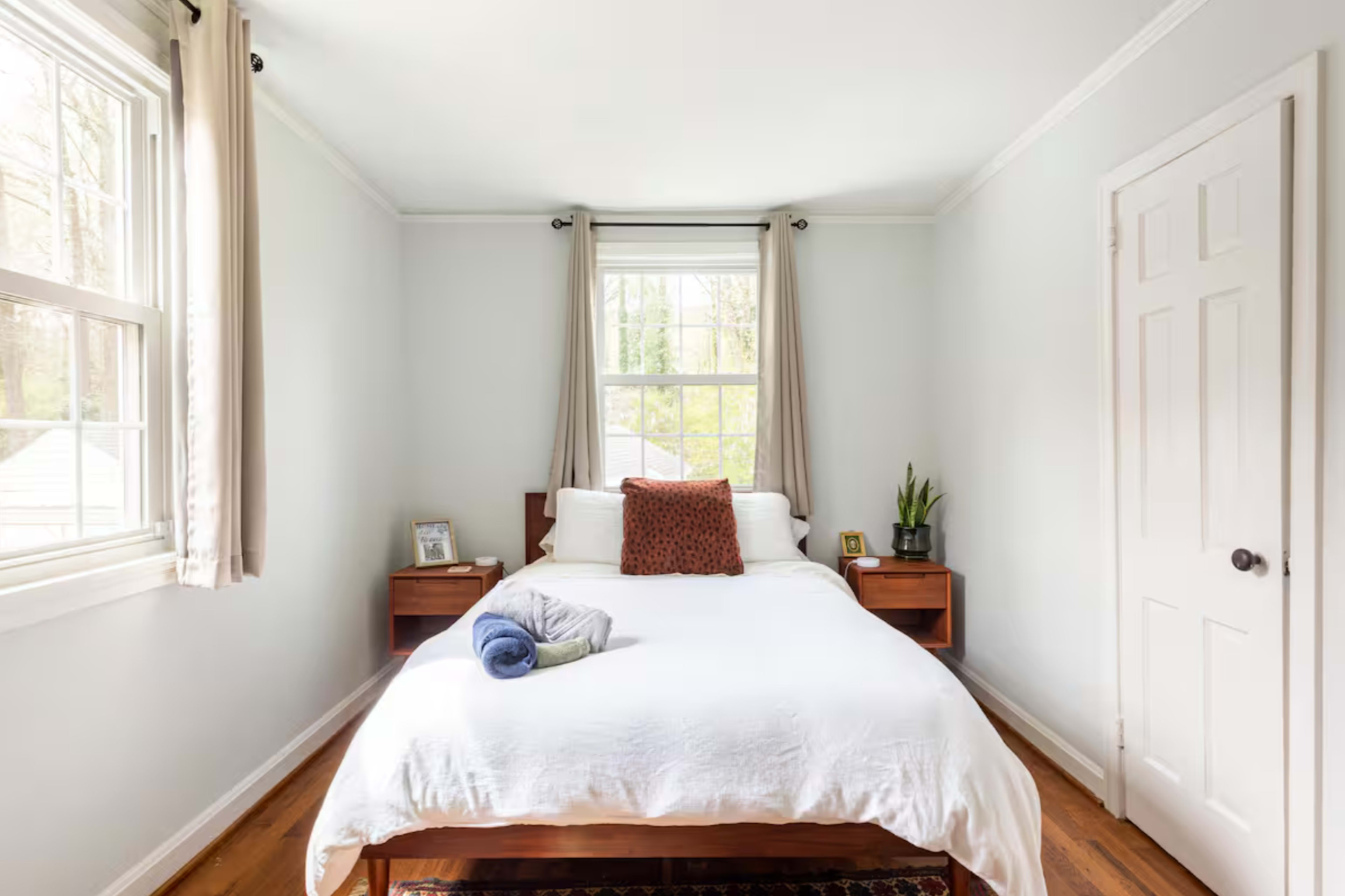 The image shows a simple, well-lit bedroom featuring a white bed with pillows and a blanket, two wooden bedside tables, and a plant on one table near a window.
