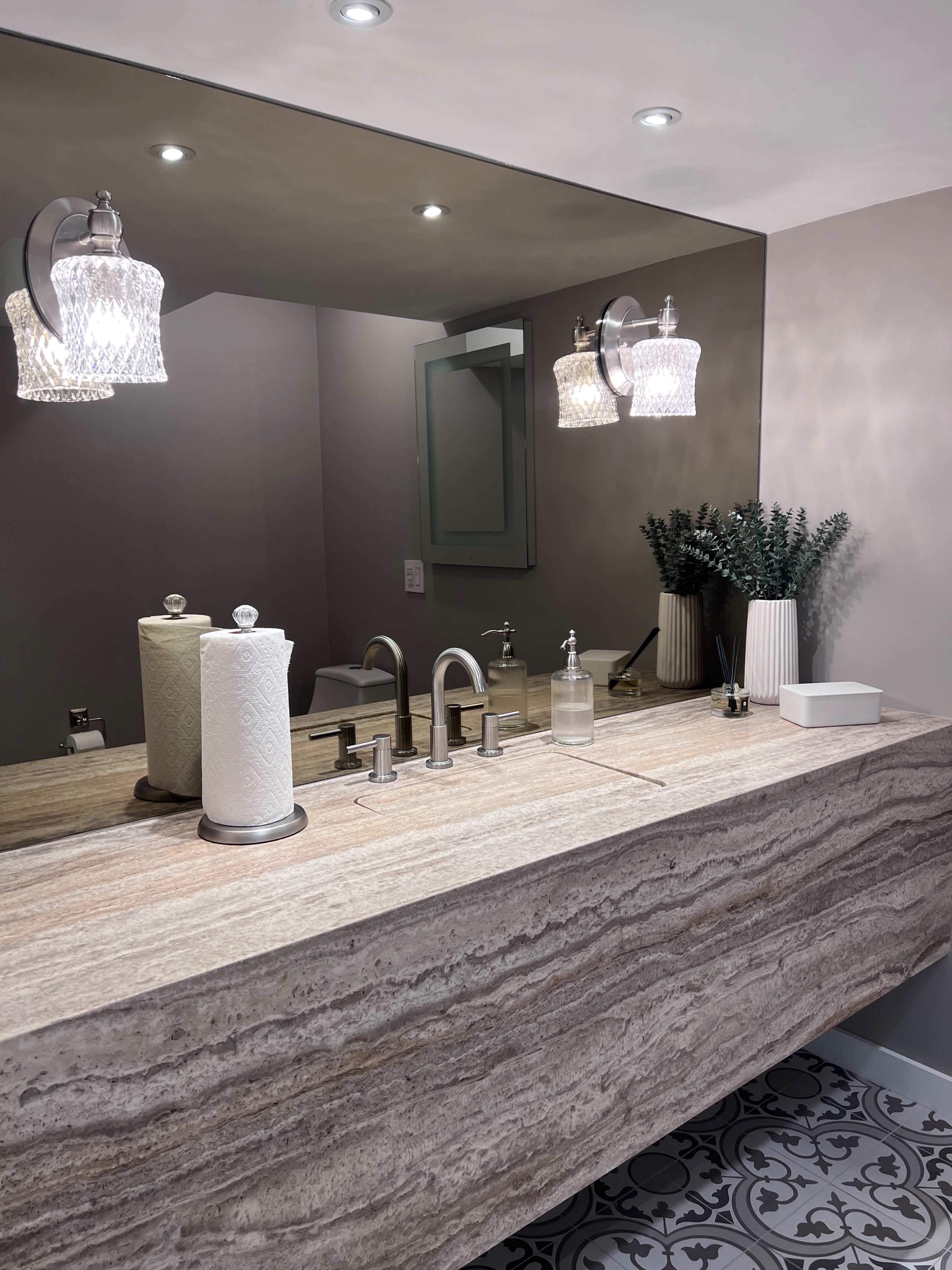 A modern bathroom features a polished stone countertop with dual faucets, a large mirror, and stylish light fixtures above.
