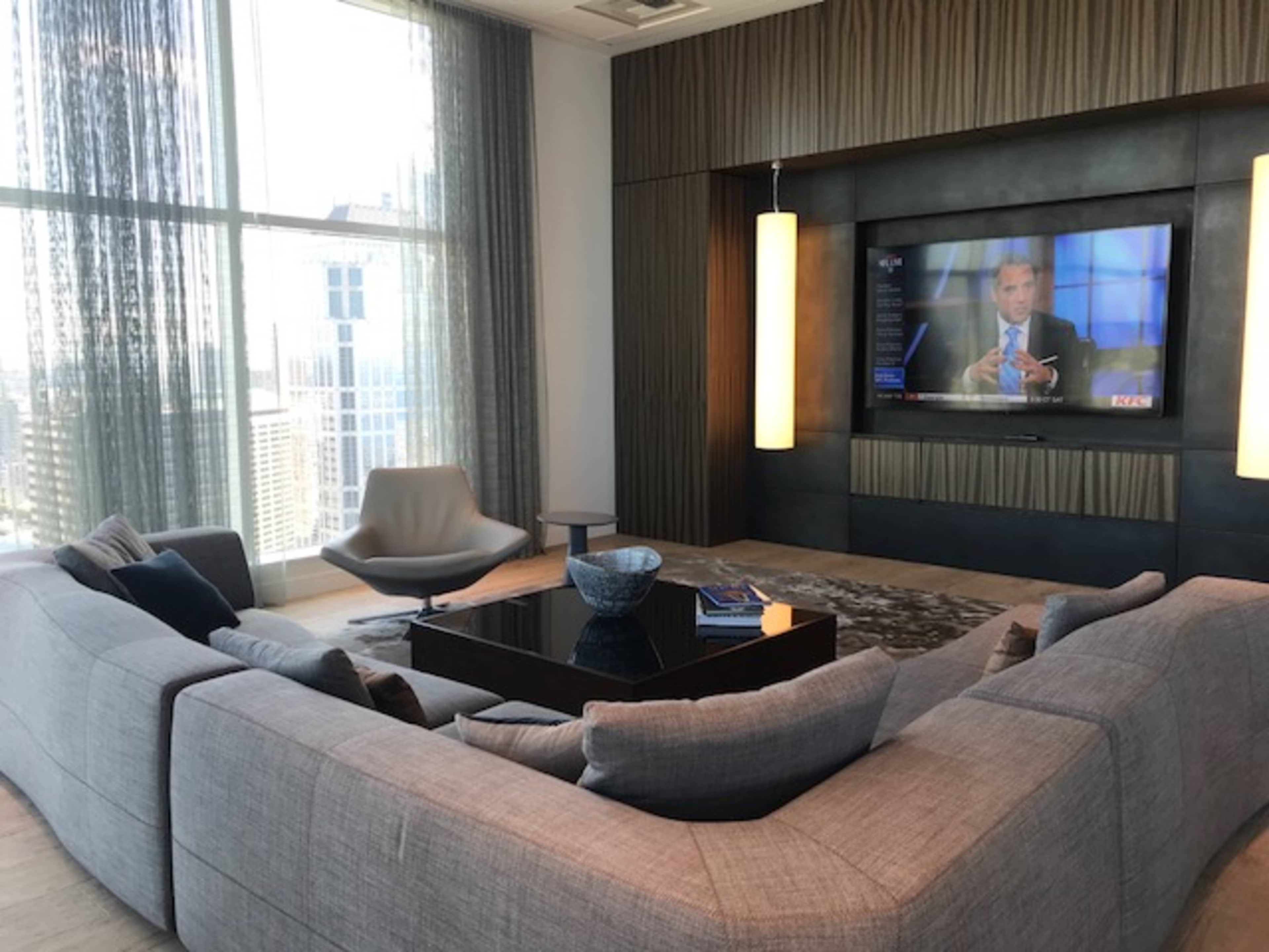 A modern living room features a large sectional sofa, a black coffee table, and a television mounted on the wall, with floor-to-ceiling windows showing a cityscape.