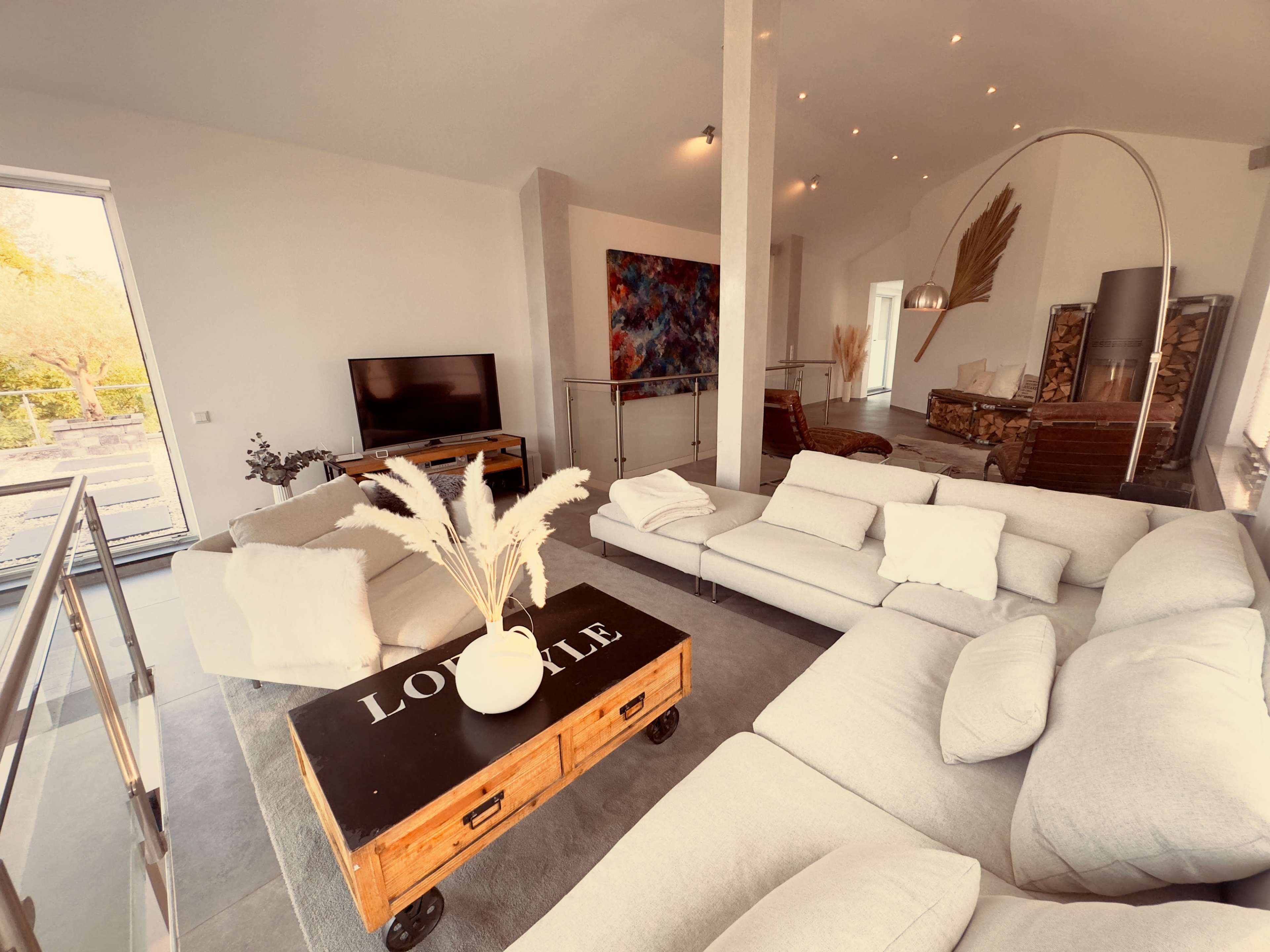 A modern living room features a large sectional sofa, a wooden coffee table, and a flat-screen TV, with large windows providing natural light.