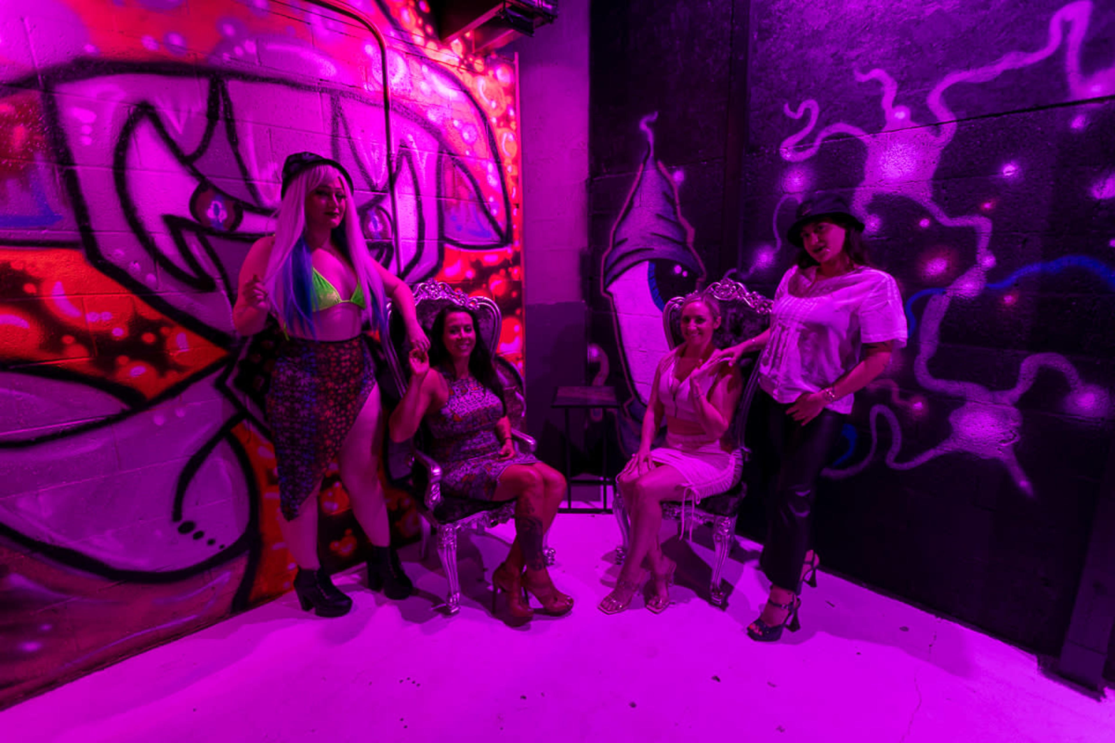 Four women sit in ornate chairs against a colorful graffiti wall, illuminated by purple lighting.