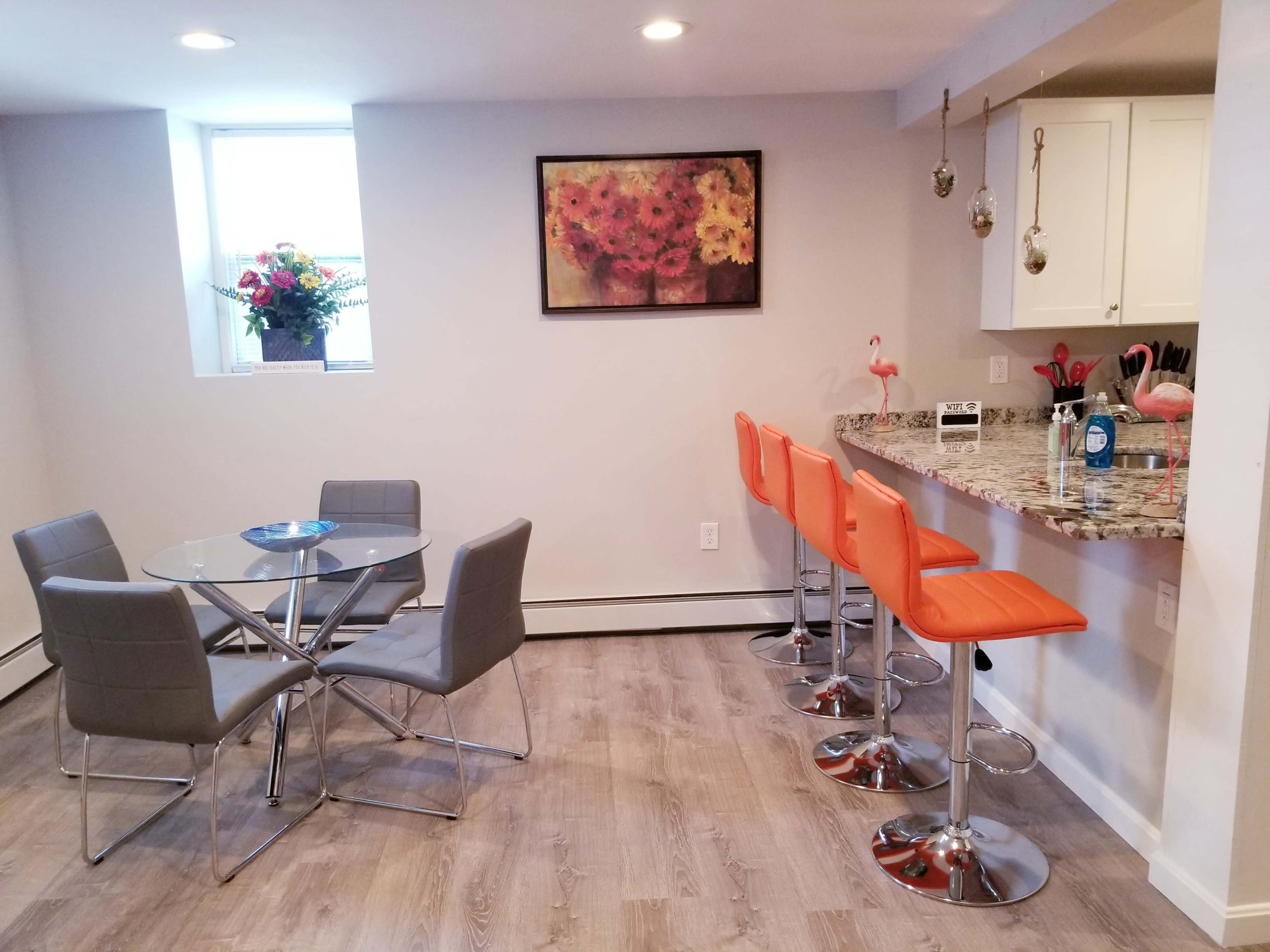 A modern kitchen and dining area featuring a circular glass table with gray chairs and a bar counter with orange stools.