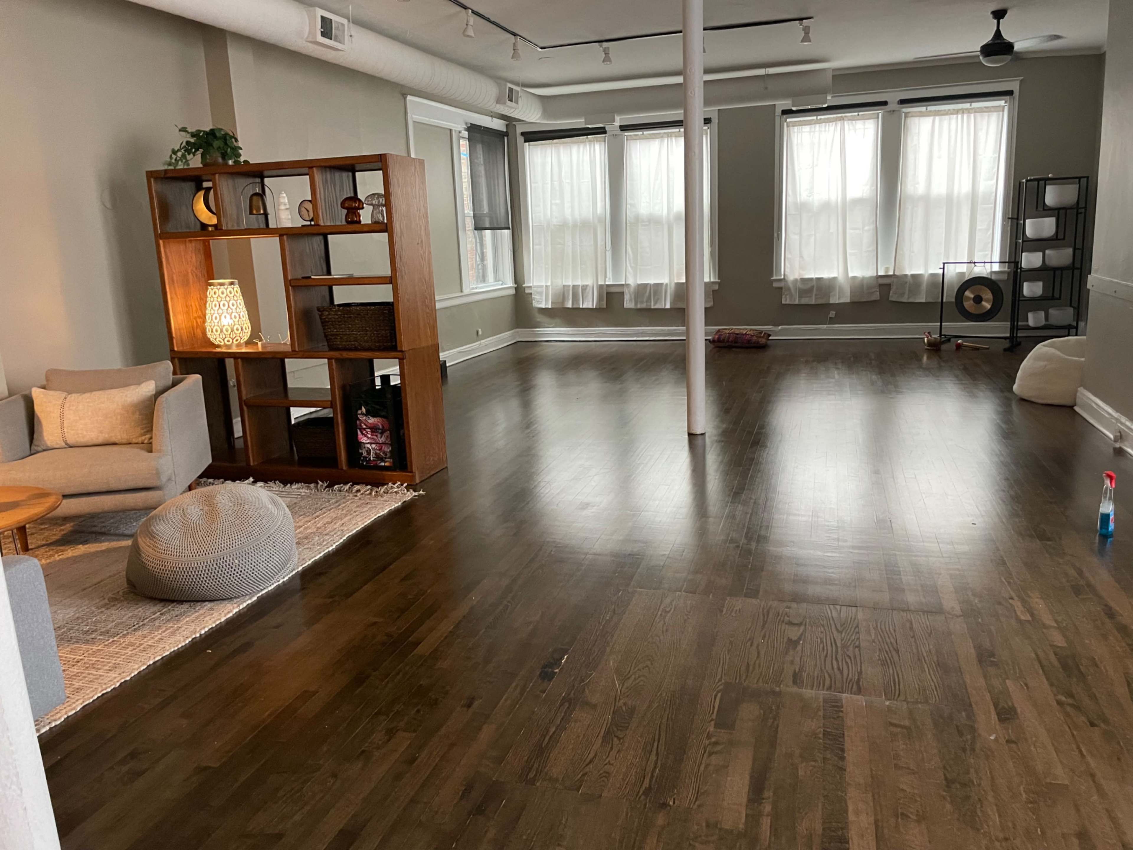 The image shows a spacious, uncluttered room with wooden floors, large windows, and minimal furniture including a shelf, a seating area, and a few decorative items.