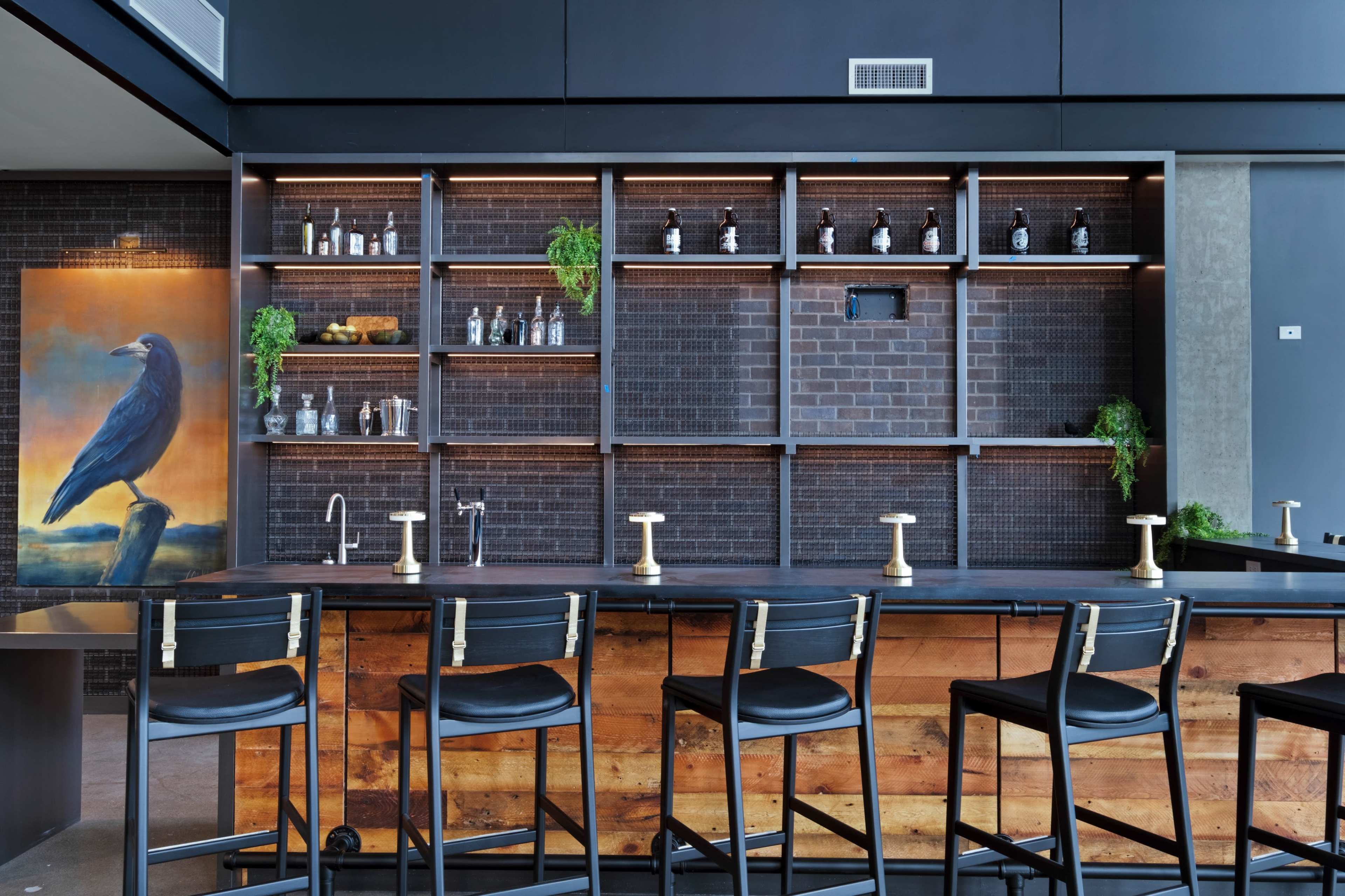 The image shows a modern bar with a wooden counter, high black stools, and a backdrop featuring shelves stocked with glassware and bottles, alongside a large painting of a bird.