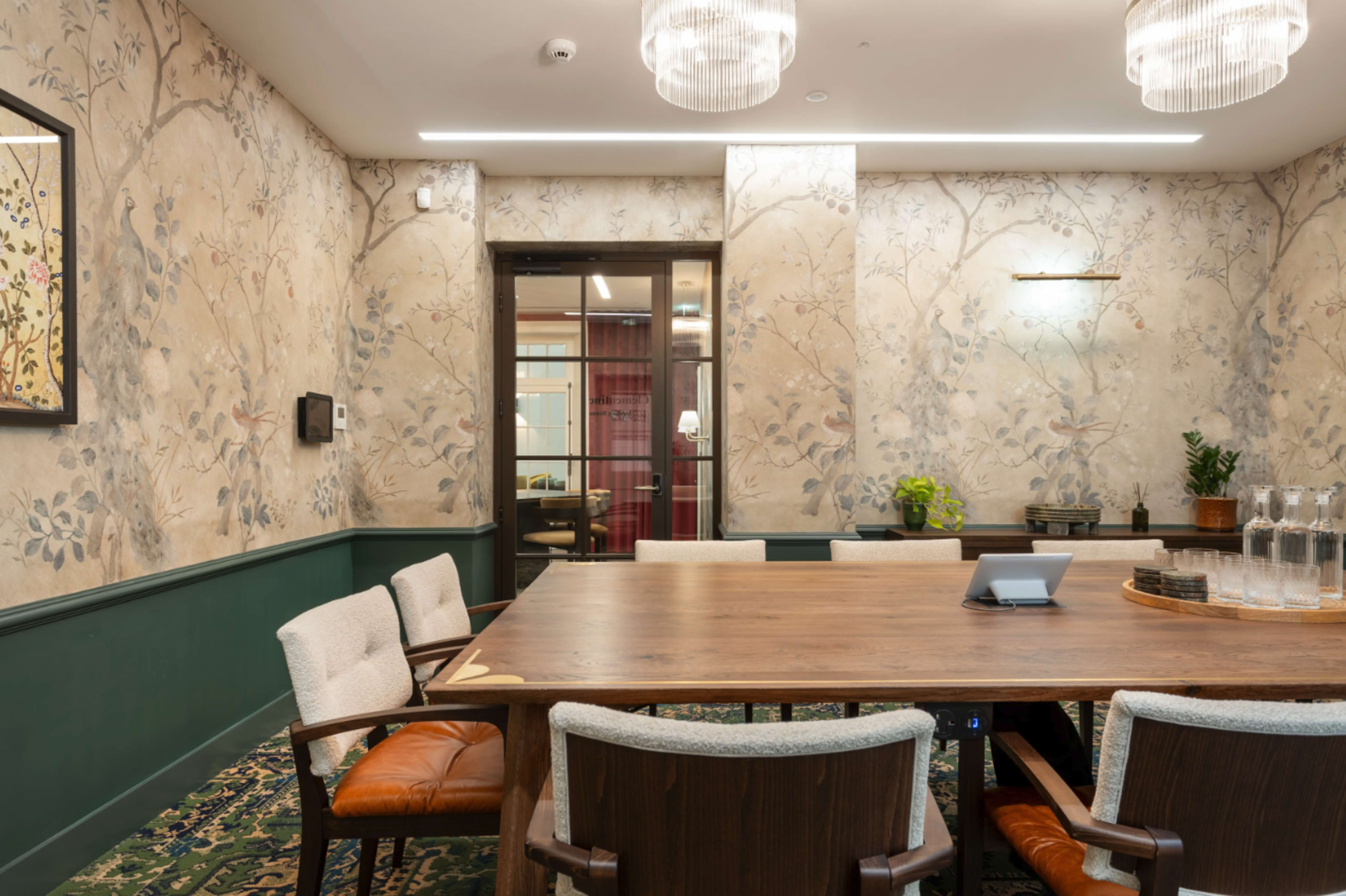 The image shows a conference room with a large wooden table surrounded by upholstered chairs, adorned with floral wallpaper and equipped with a glass door leading to another room.