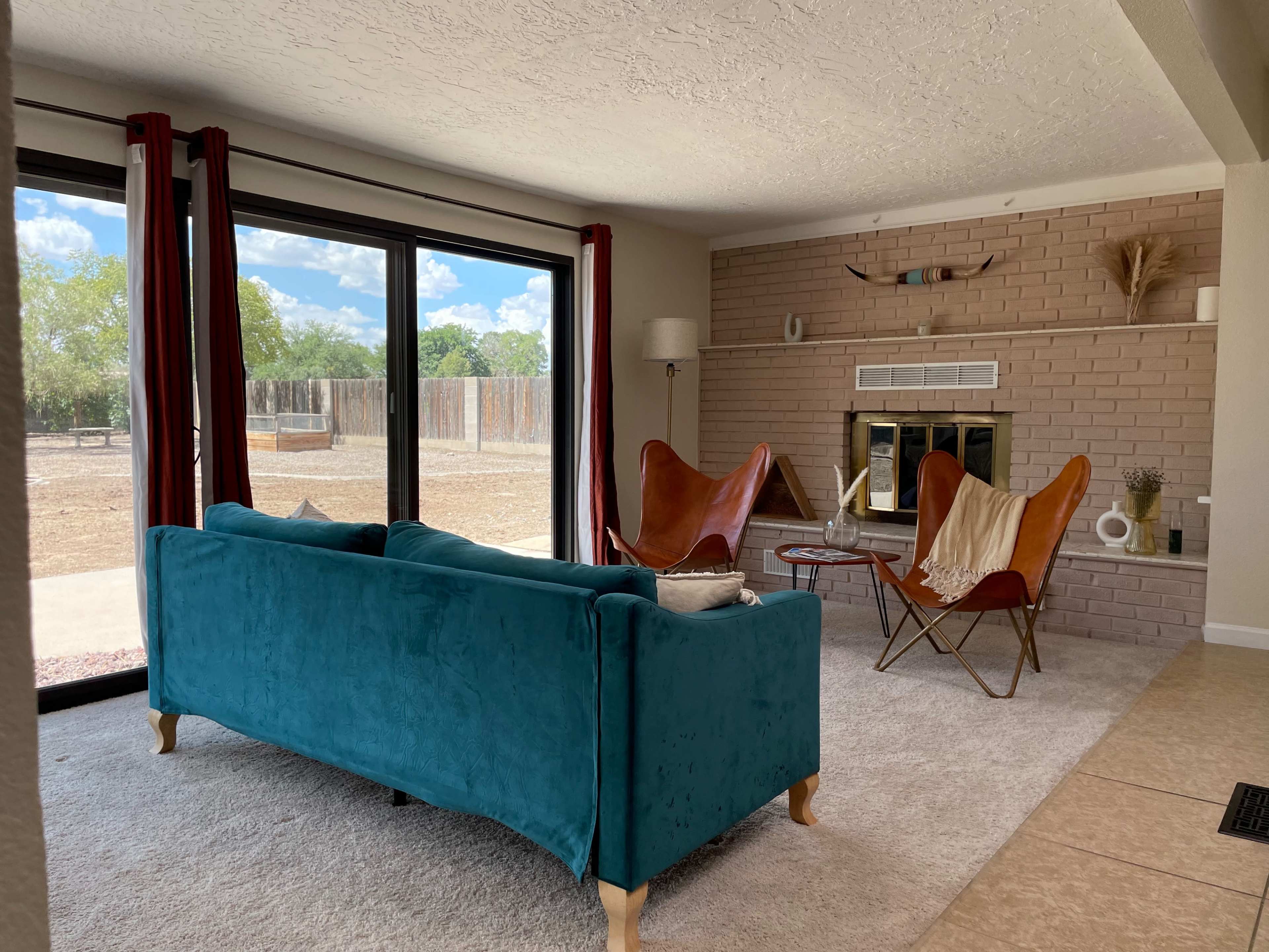 A modern living room with a turquoise sofa, two leather butterfly chairs, and a stone fireplace, featuring large windows that overlook a backyard.