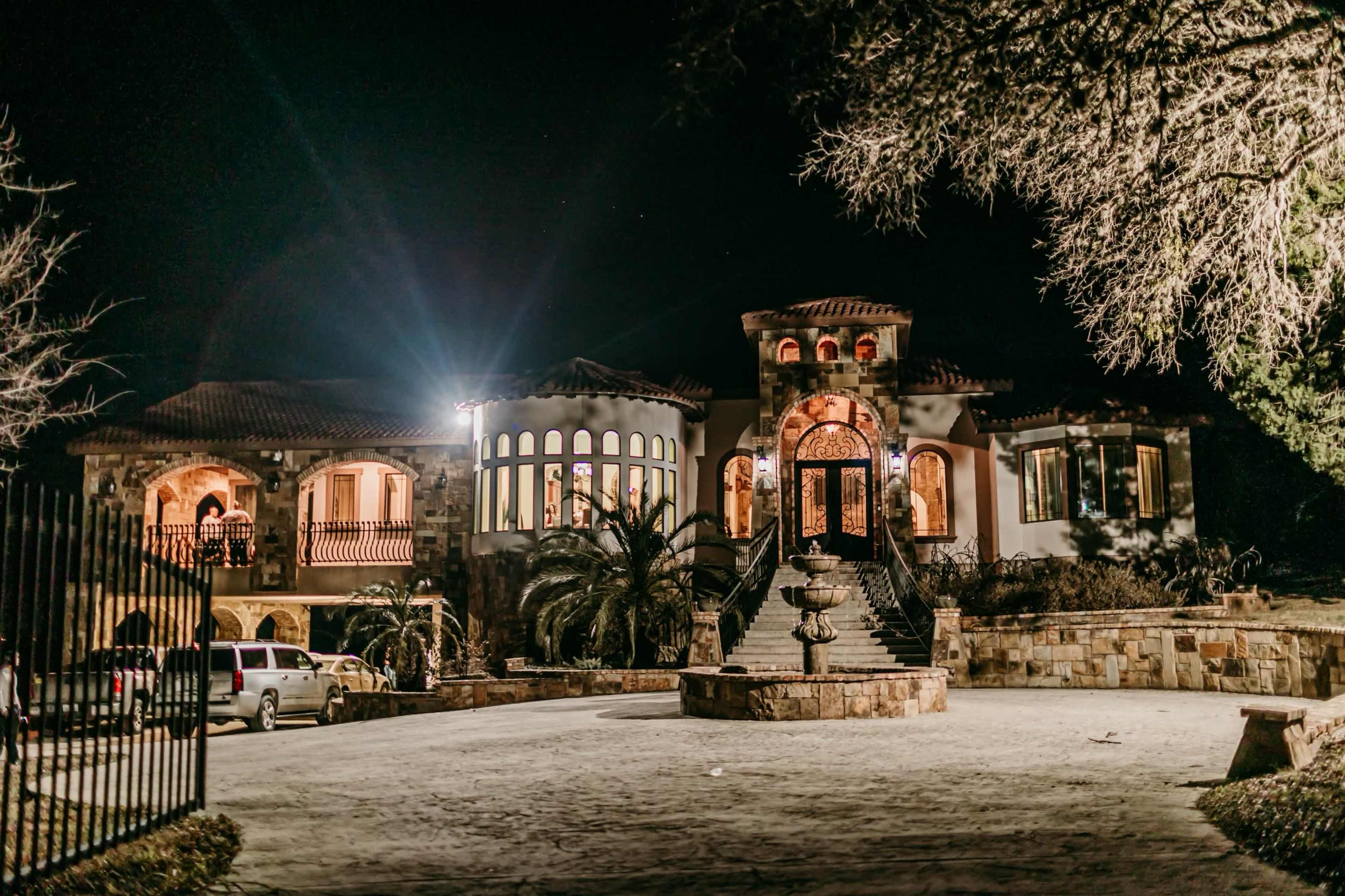 A large, intricately designed mansion with a well-lit facade and a fountain in the front yard is shown at night.