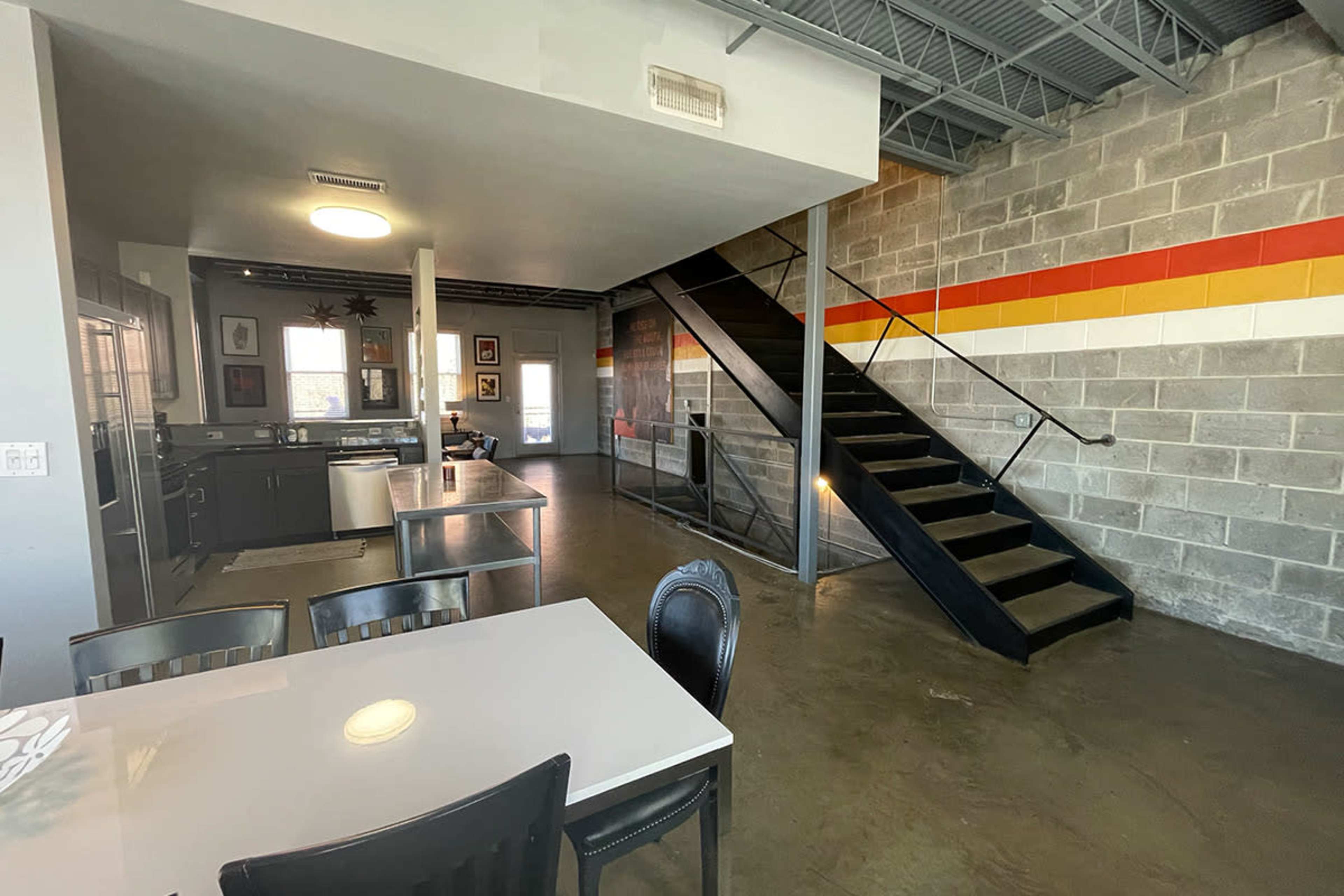 The image shows an industrial-style interior featuring an open floor plan with a dining table, kitchen, and a staircase leading to an upper level.