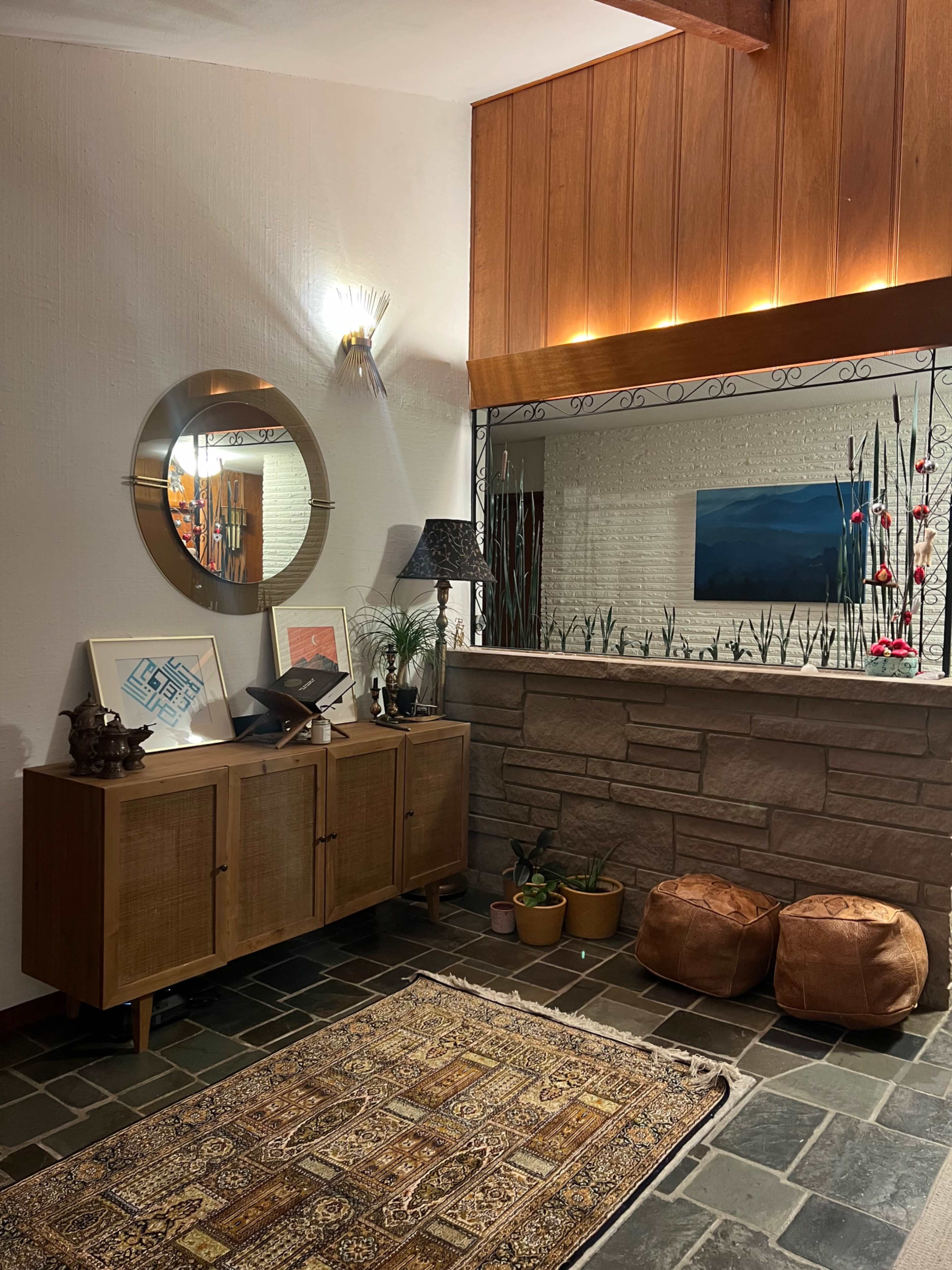 A cozy entryway featuring a wooden console table with decorative items, a round mirror, and a stone wall with potted plants and floor cushions.