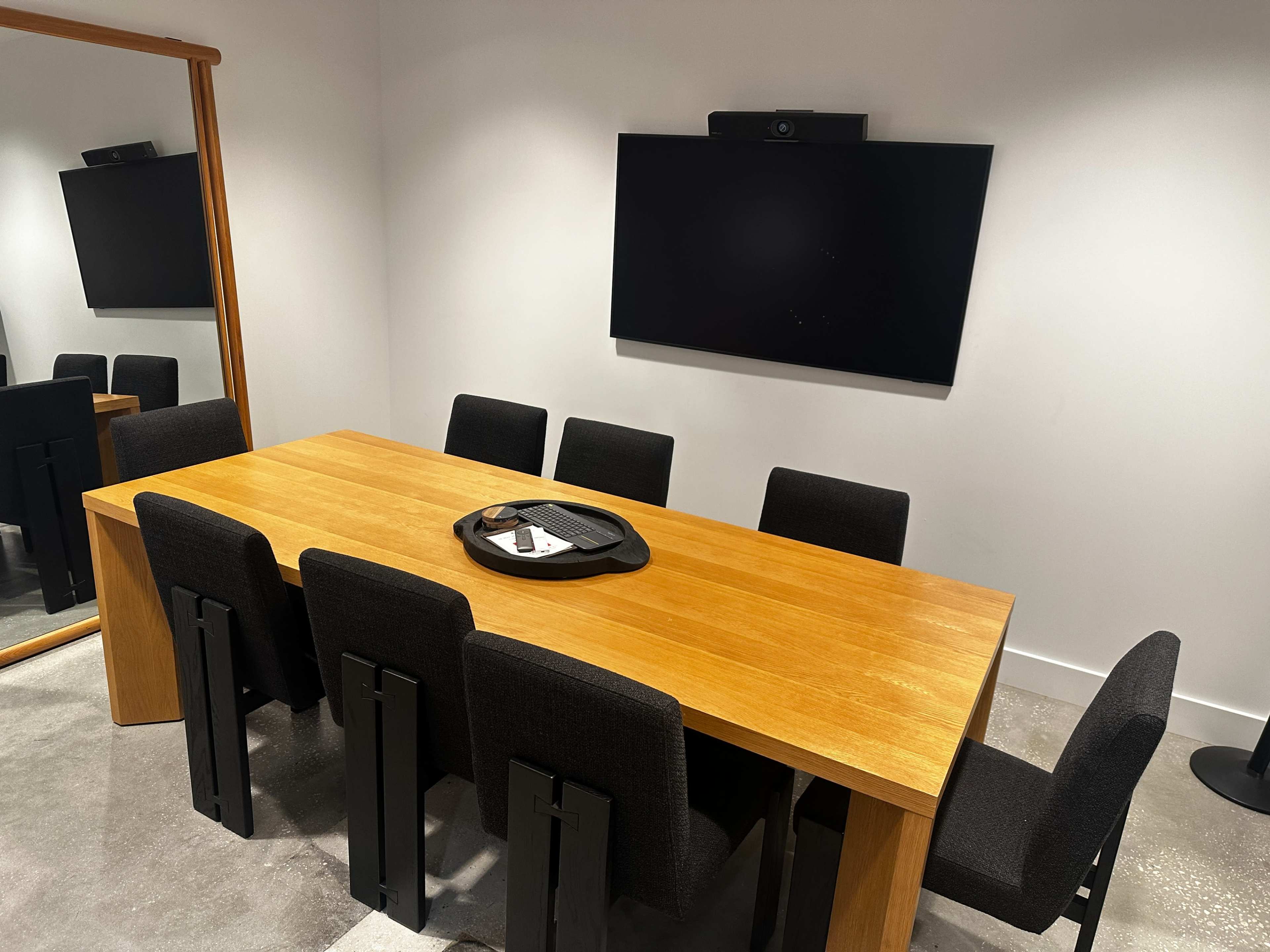 The image shows a conference room with a large wooden table surrounded by black upholstered chairs, a mounted screen on the wall, and a mirror reflecting the room.