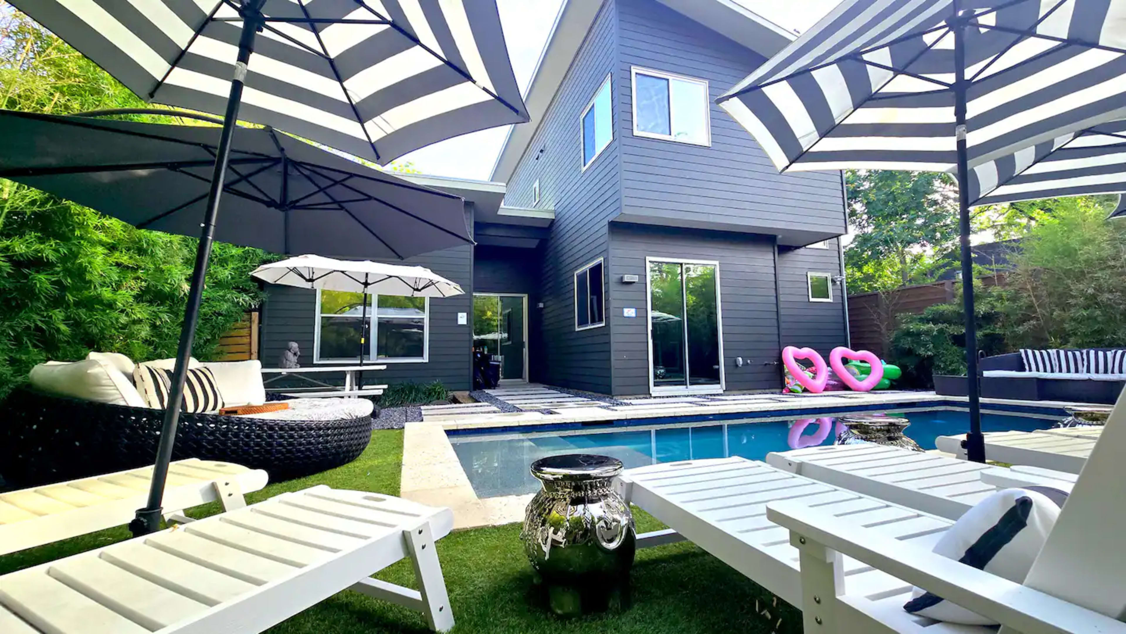 The image depicts a modern backyard with a swimming pool surrounded by lounge chairs, umbrellas, and a two-story dark-colored house in the background.