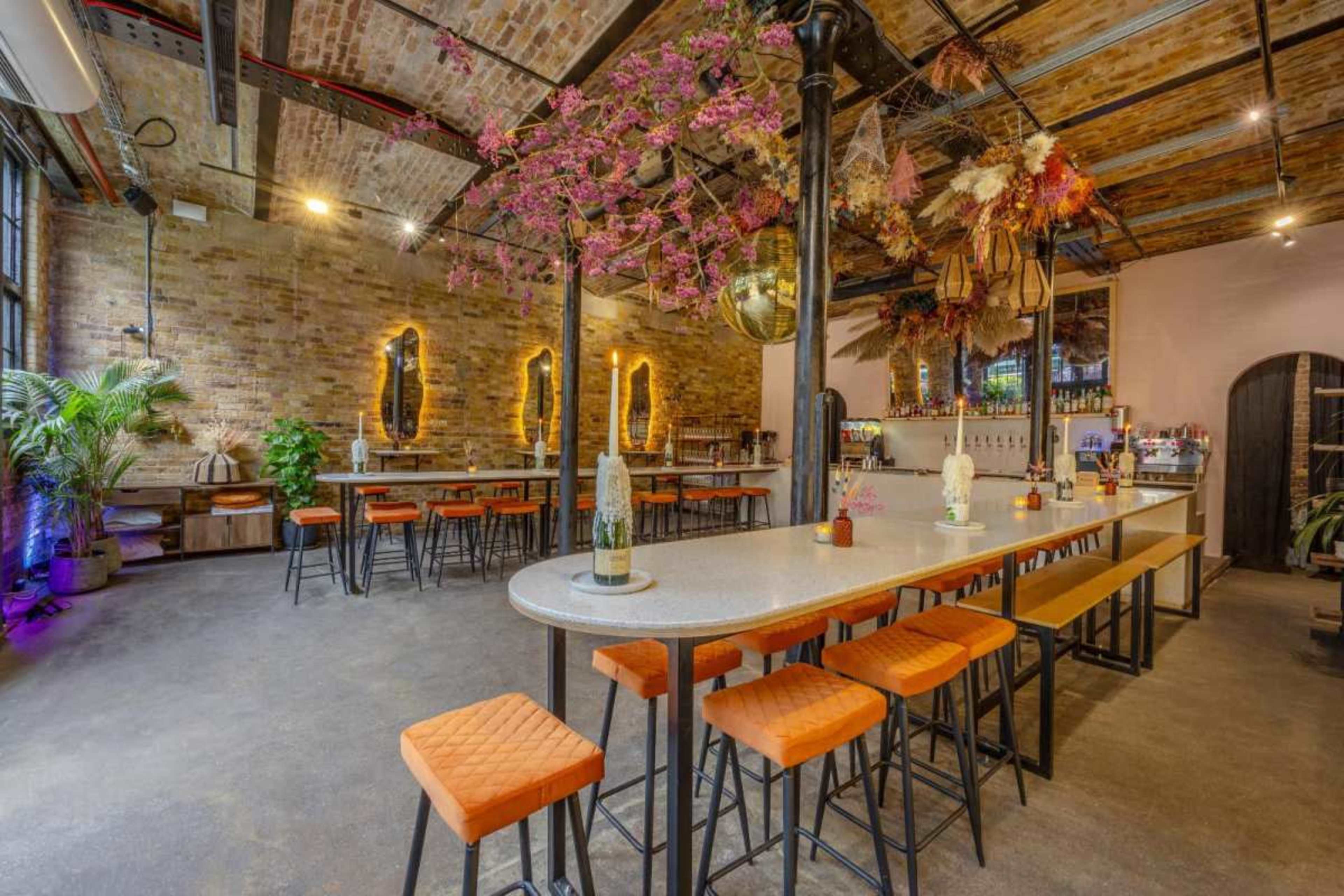 A spacious, modern bar area with long tables and orange stools, complemented by decorative plants and hanging floral arrangements.