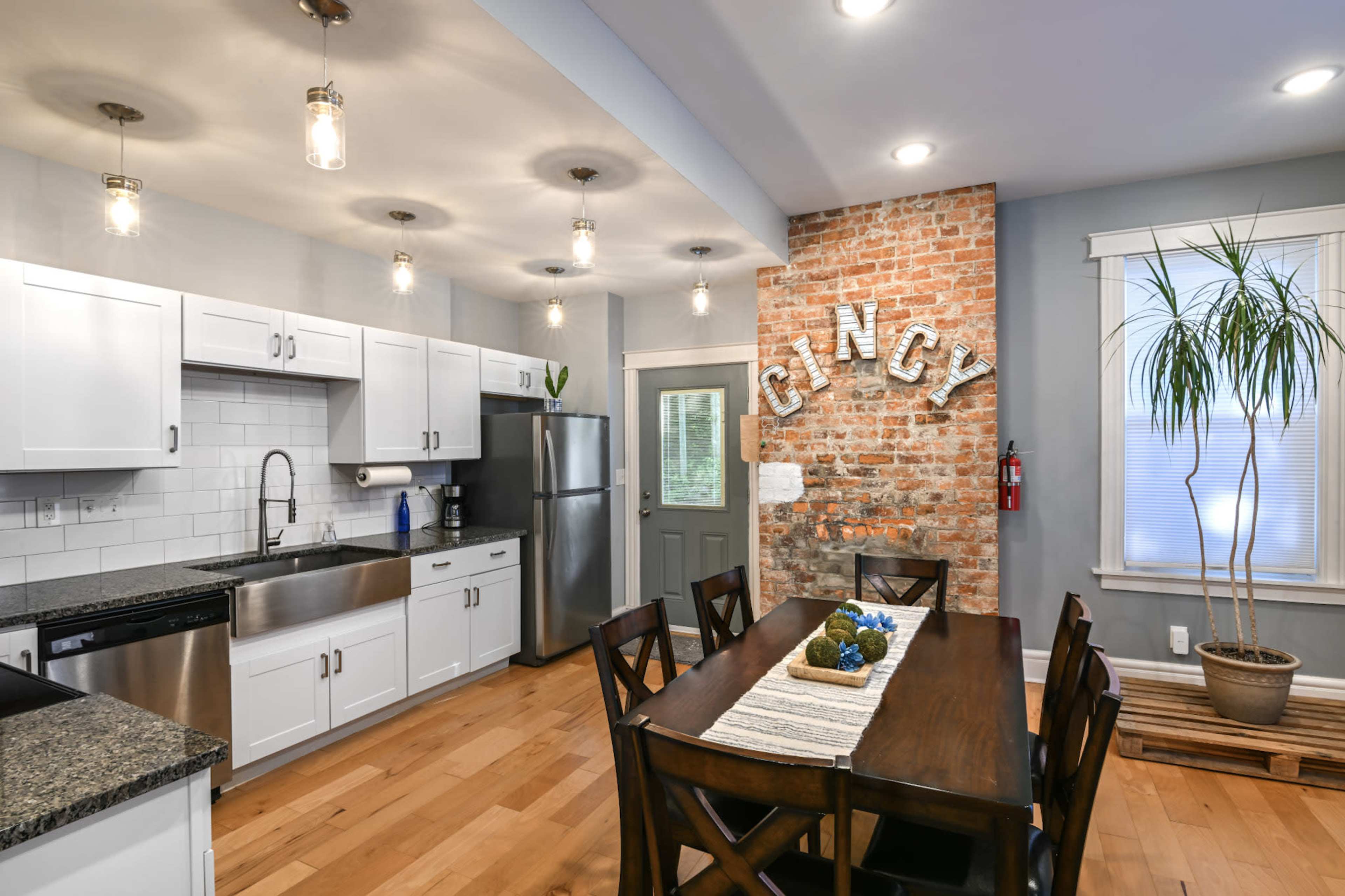 The kitchen features white cabinets, stainless steel appliances, and a wooden dining table situated under exposed bulb lighting, with a brick accent wall displaying the word "CINCY."