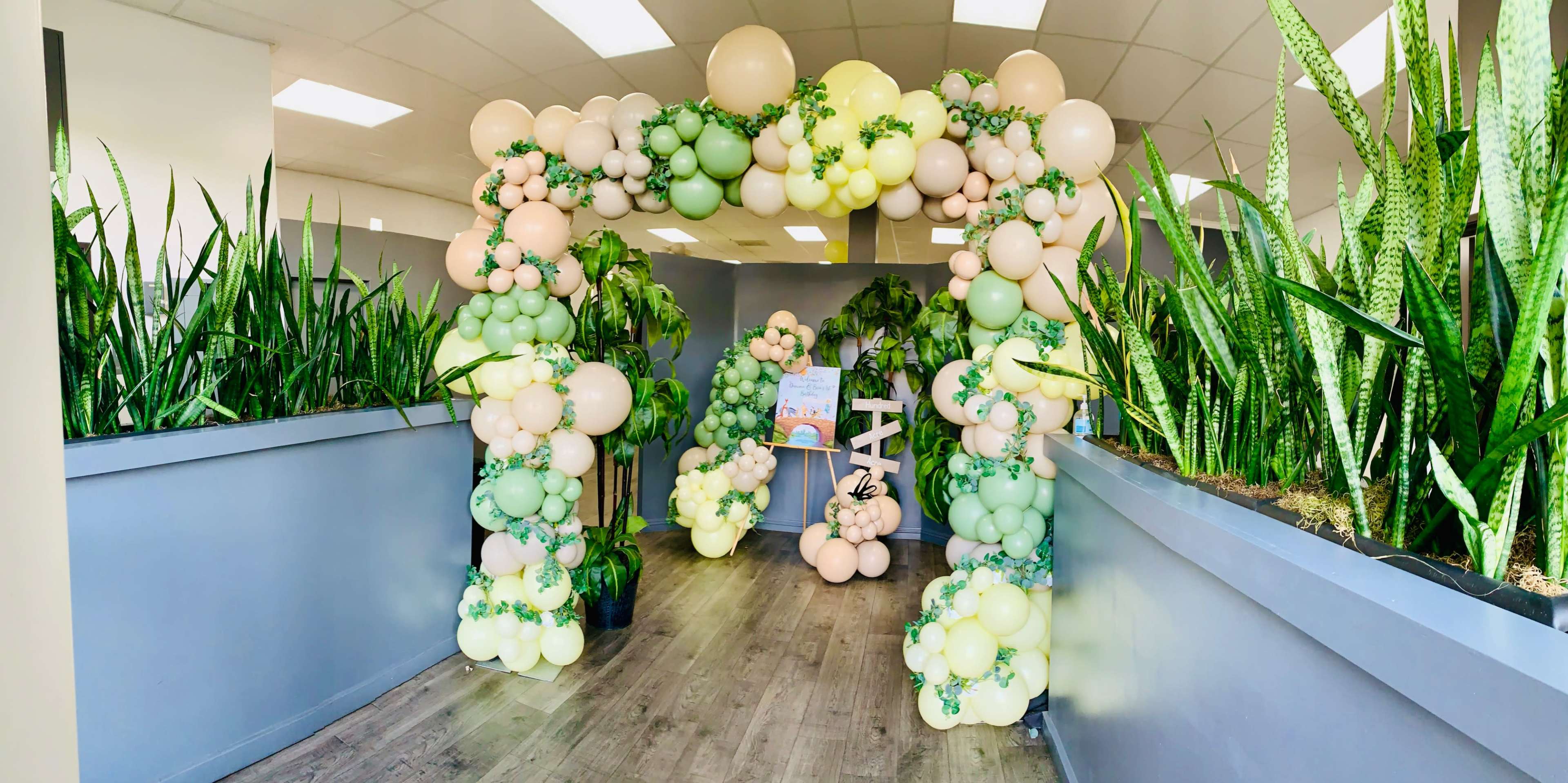 A large balloon arch in pastel colors is set up in a hallway surrounded by tall green plants.