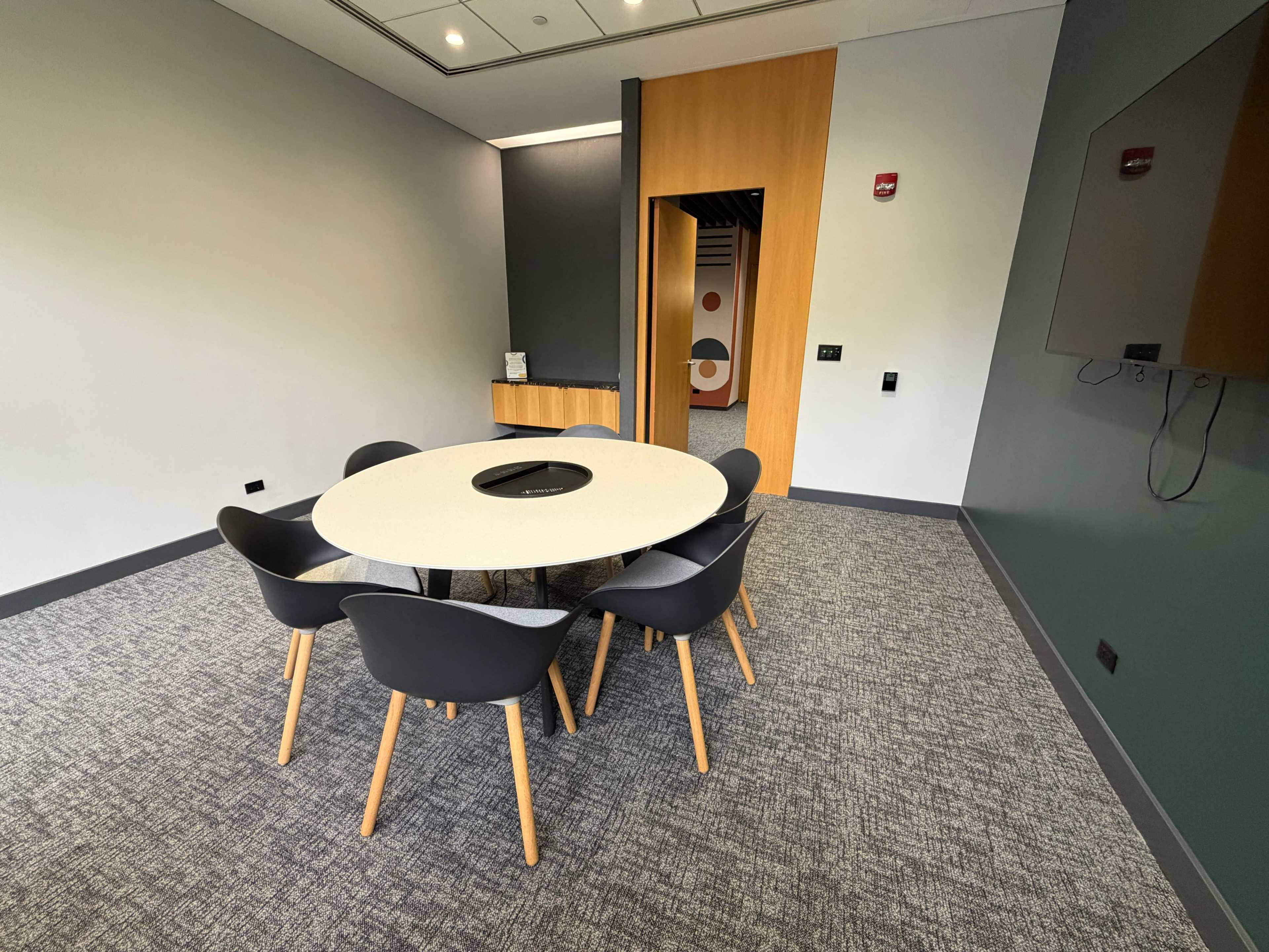 A modern conference room features a round table surrounded by six chairs, with a minimalist design and a large screen on the wall.