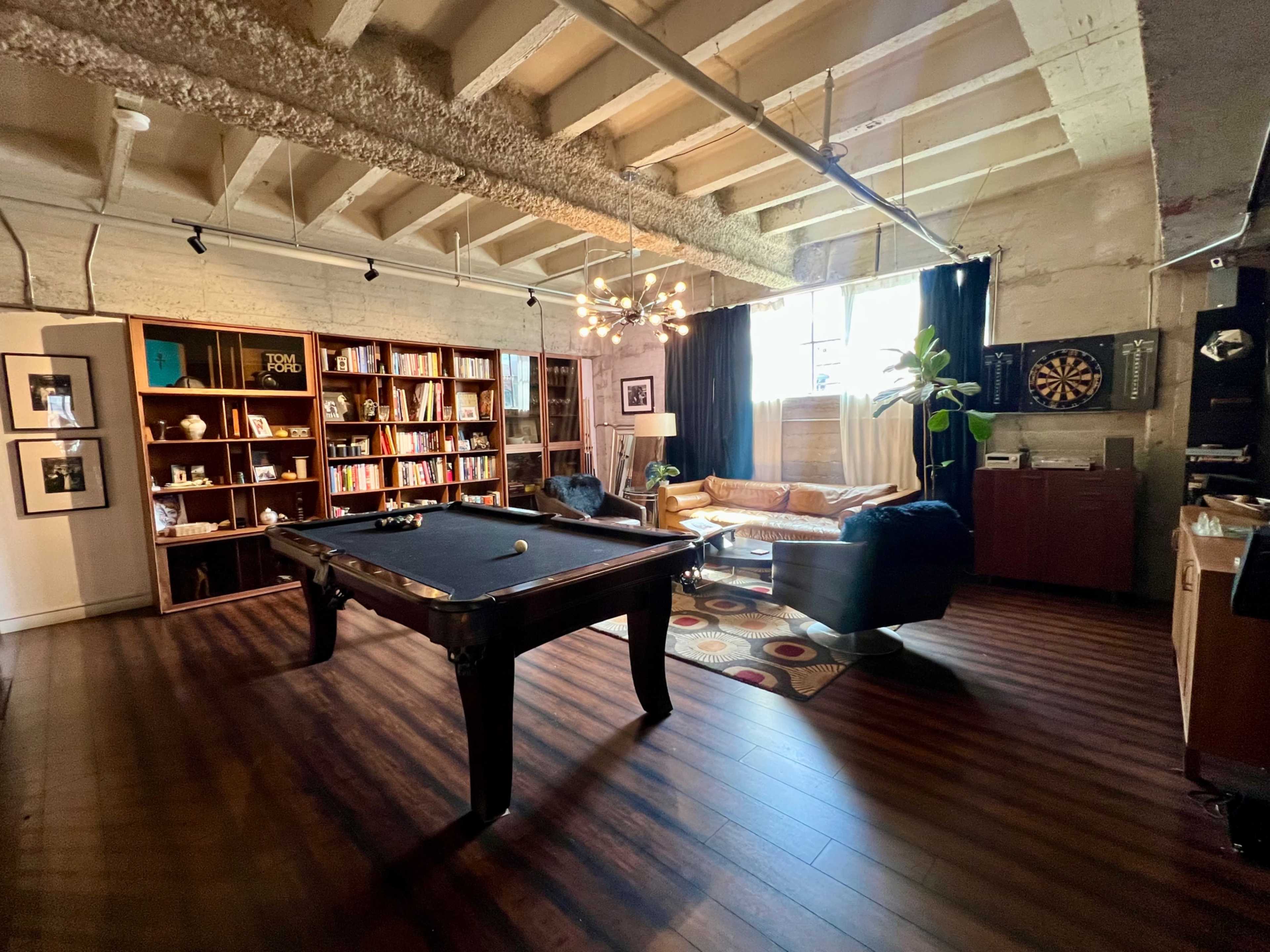 The image shows a spacious interior that features a pool table, a bookshelf filled with books, a seating area with a couch, and various decorative elements, illuminated by natural light coming through a window.
