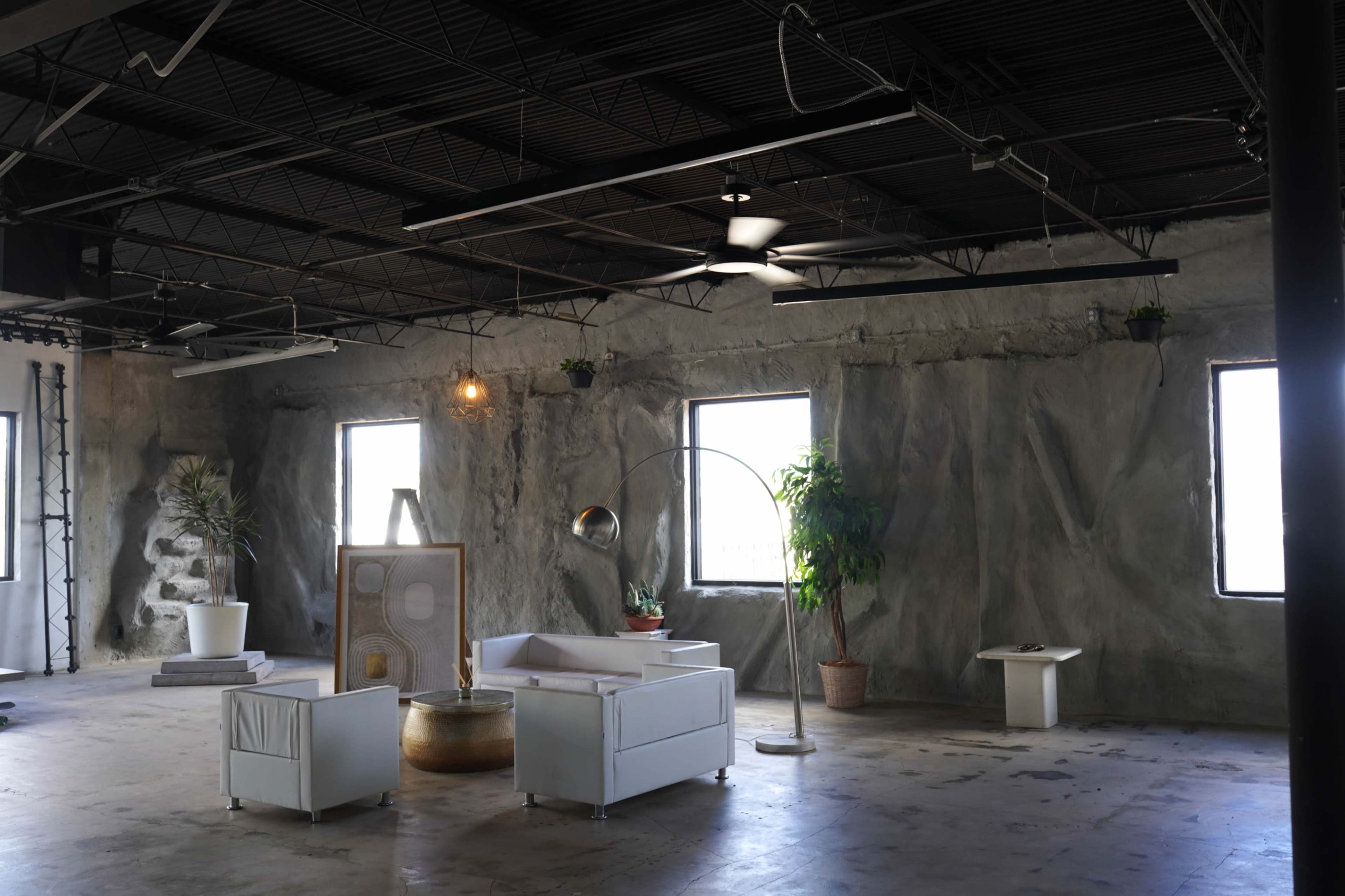 The image shows a spacious, industrial-style room with gray walls, large windows, and minimalist white furniture, including a sofa and a lamp.