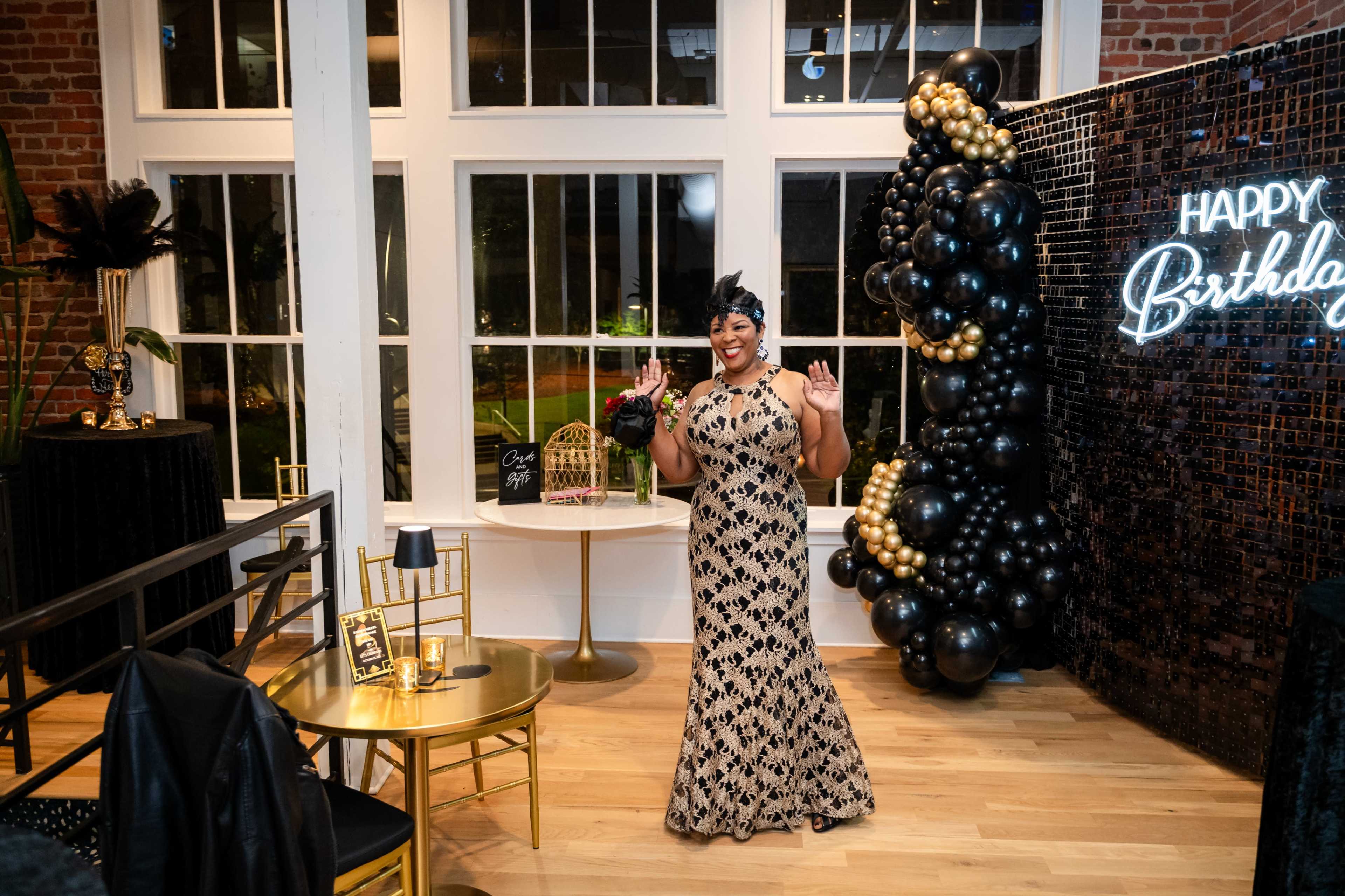 A woman in a detailed gown stands smiling in a decorated event space with a "Happy Birthday" sign and a backdrop of black and gold balloons.