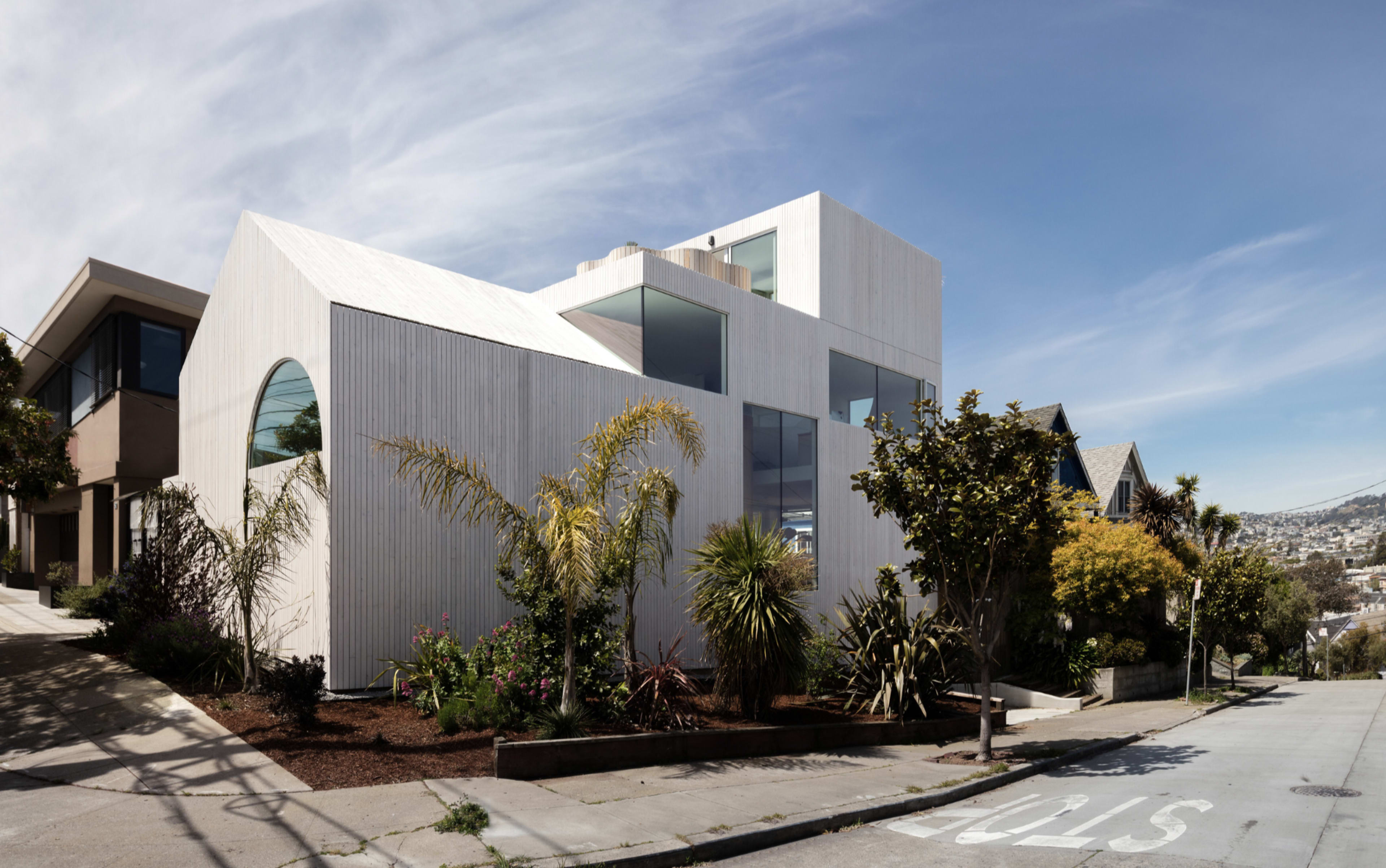 A modern, angular house with a white wood exterior and large windows is located on a sloped street, surrounded by landscaped greenery and other residential buildings.