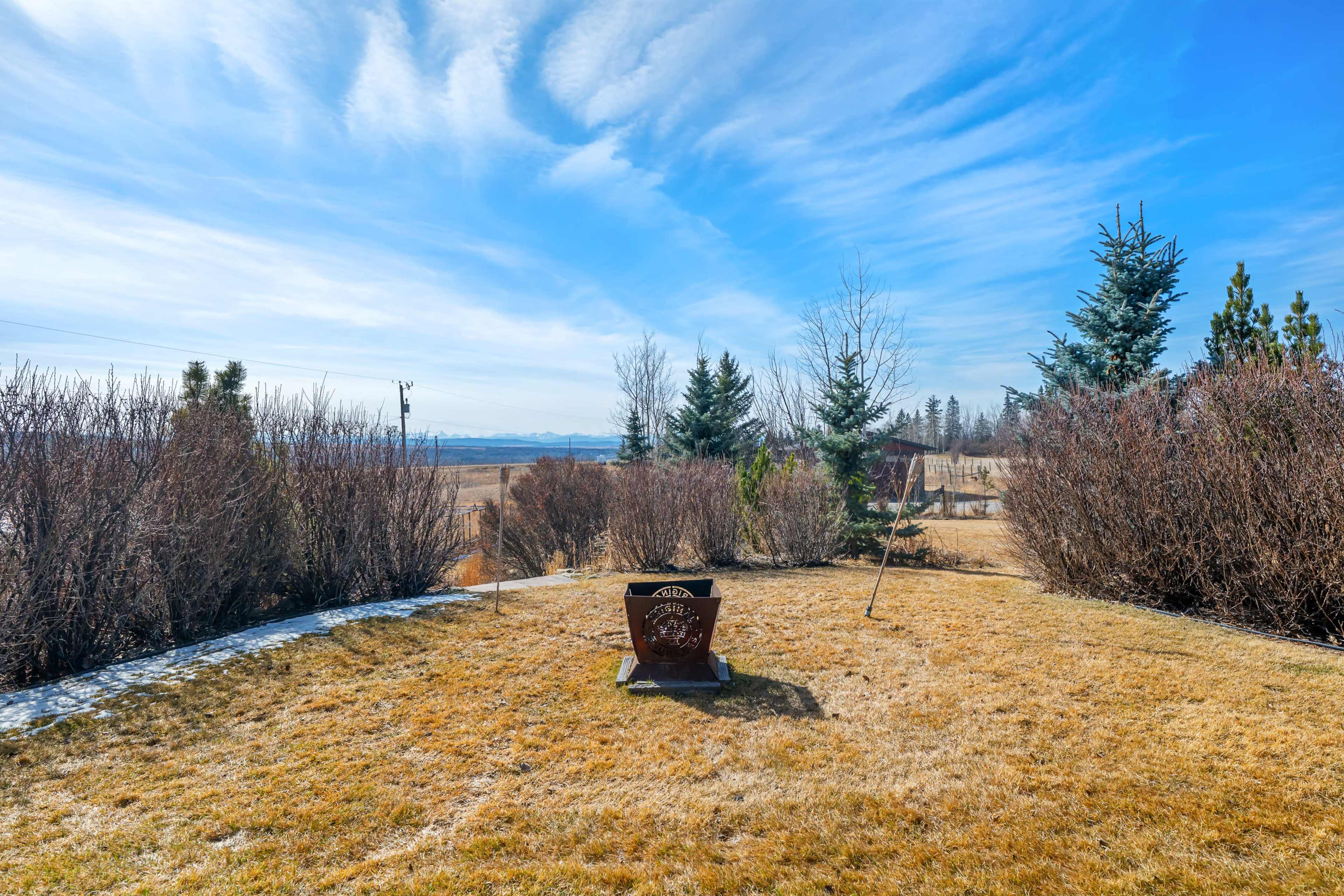 The image shows a grassy yard with a fire pit surrounded by sparse shrubs and a clear blue sky.