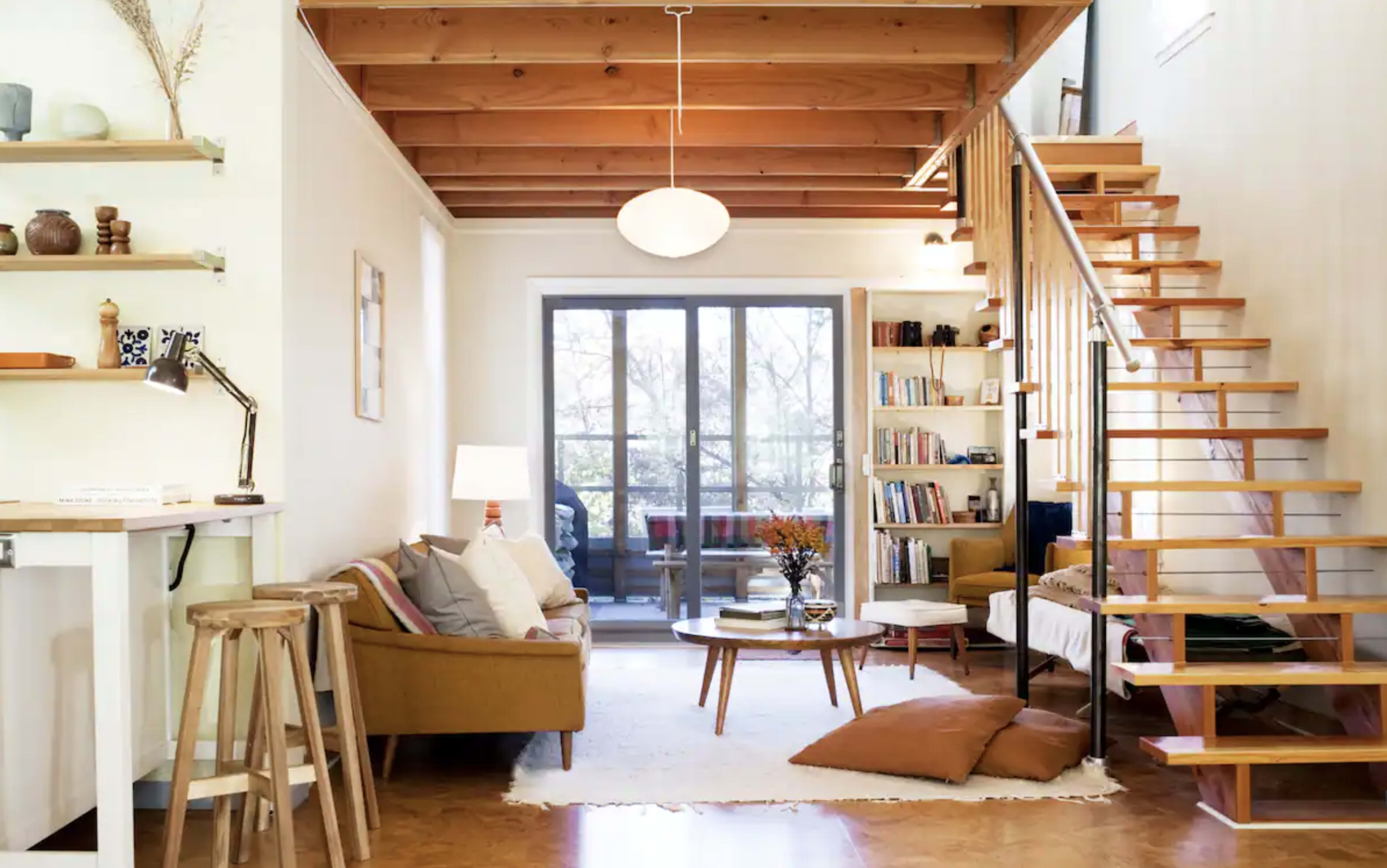 A modern living room with a wooden staircase, large windows, and a mix of seating arrangements including a sofa and armchairs.