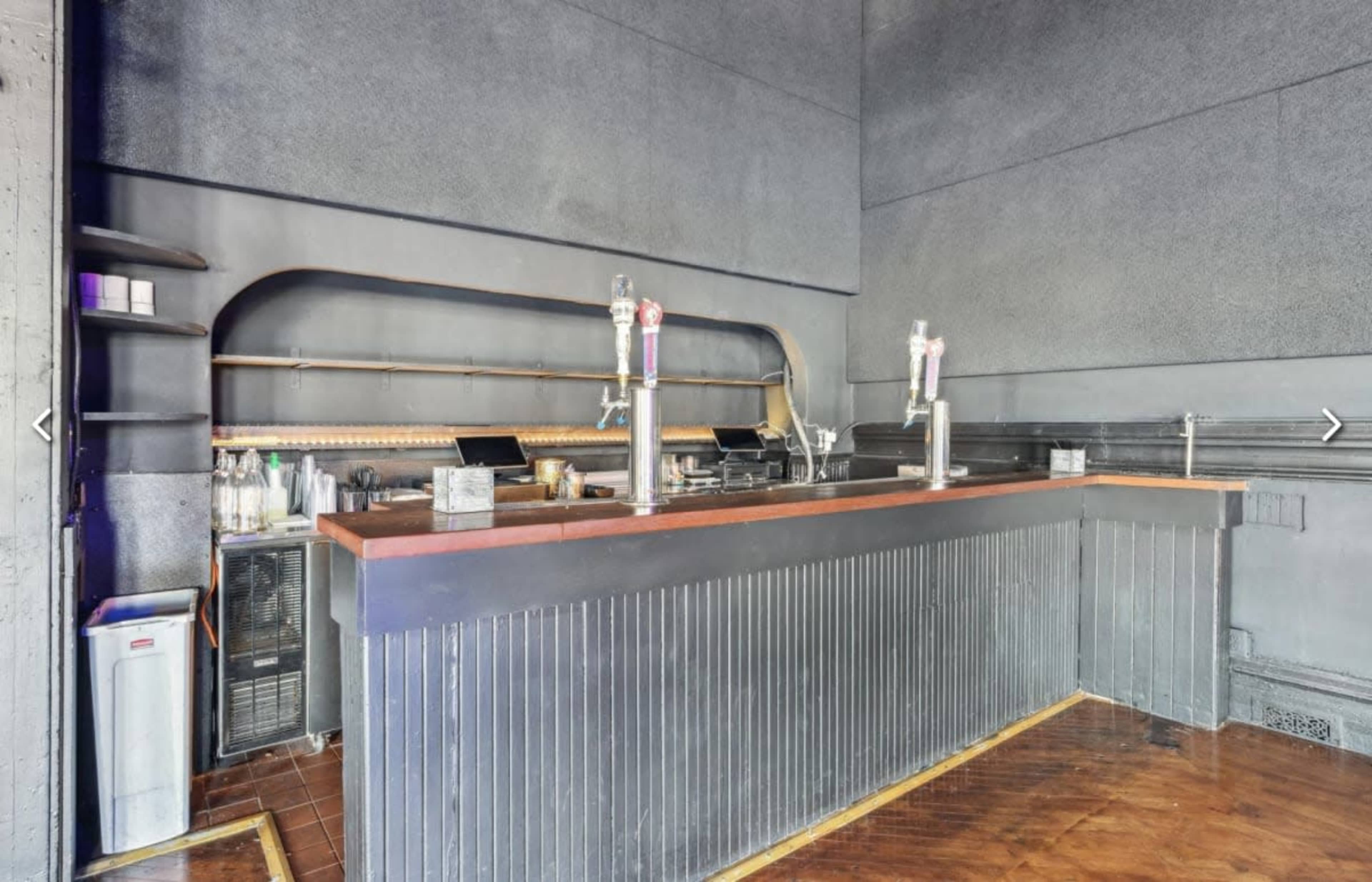 The image shows a bar area with a wooden countertop, two beer taps, and shelves for drinks against a dark wall.