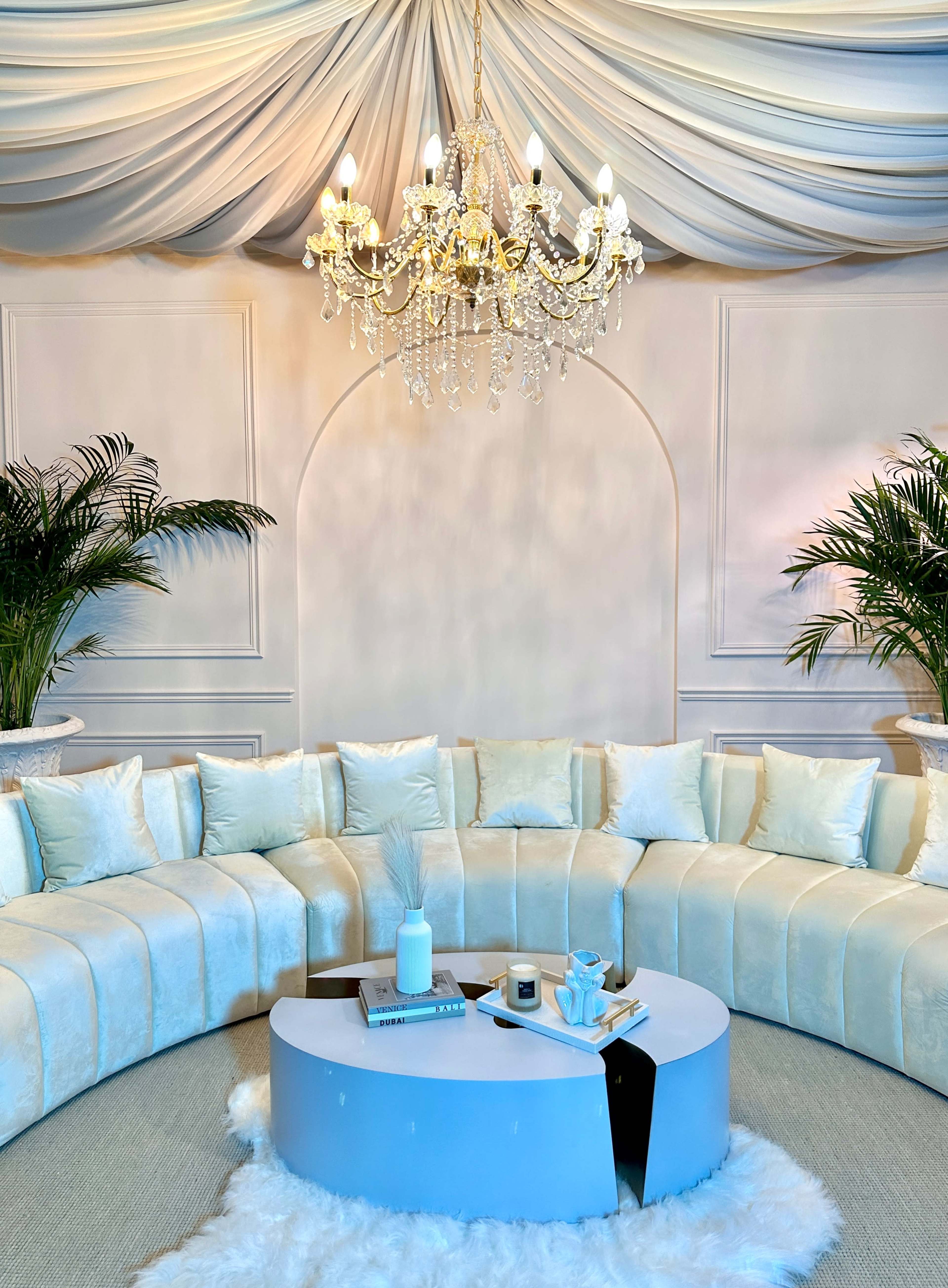 The image depicts a circular, plush seating arrangement surrounding a modern, white coffee table under a decorative chandelier, set in a stylish indoor space with draped fabric and potted plants.