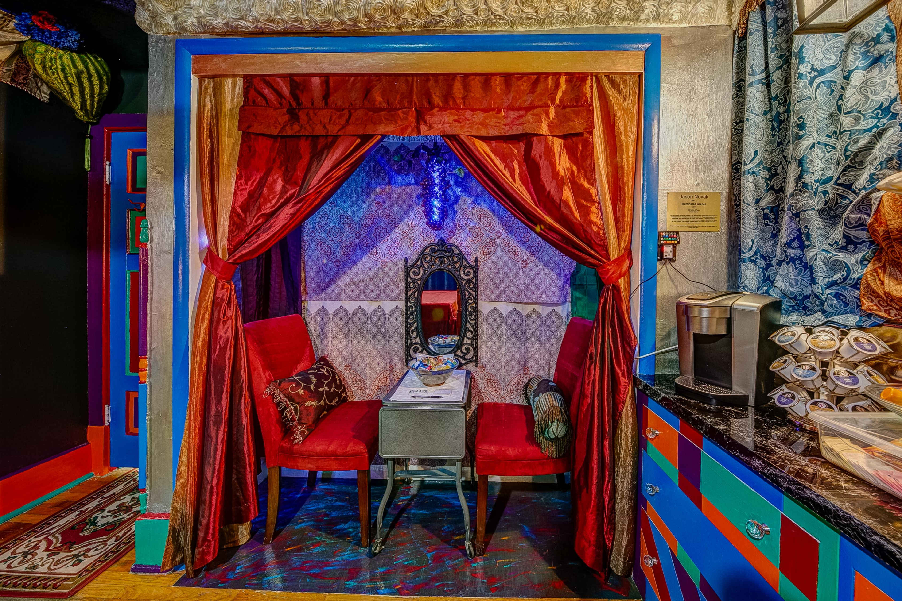 The image shows a small, cozy dining nook enclosed by red velvet curtains, featuring two red chairs and a table with a decorative mirror in a vibrant, colorfully adorned setting.