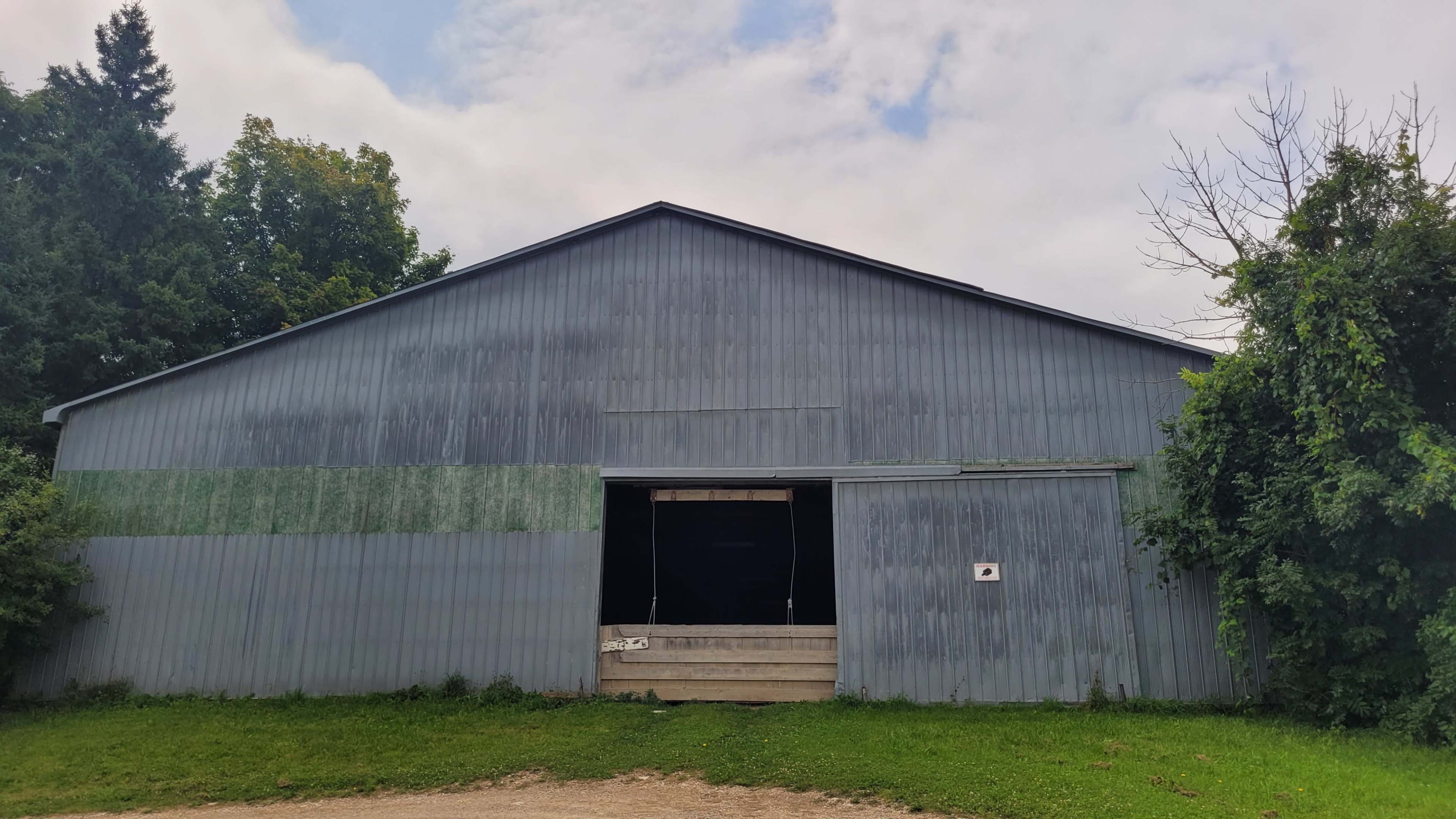 A large, gray metal barn with a partially open sliding door and a grassy area in front.