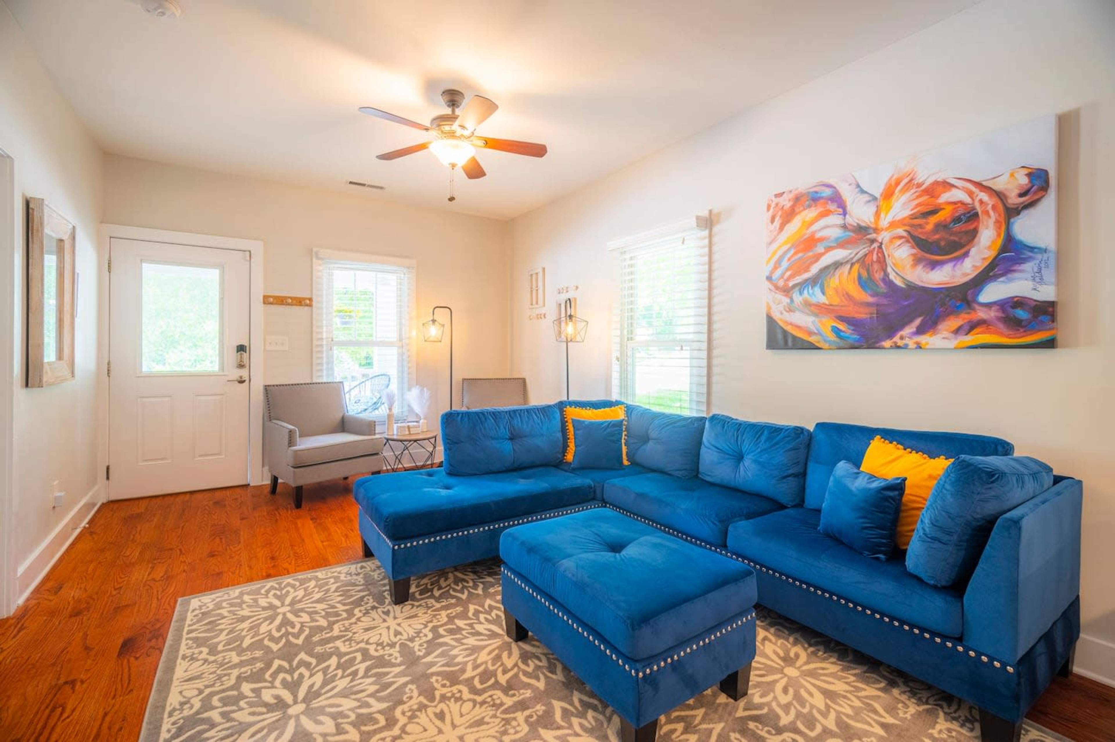 The living room features a blue sectional couch with decorative cushions, a ceiling fan, an area rug, and a colorful piece of wall art.