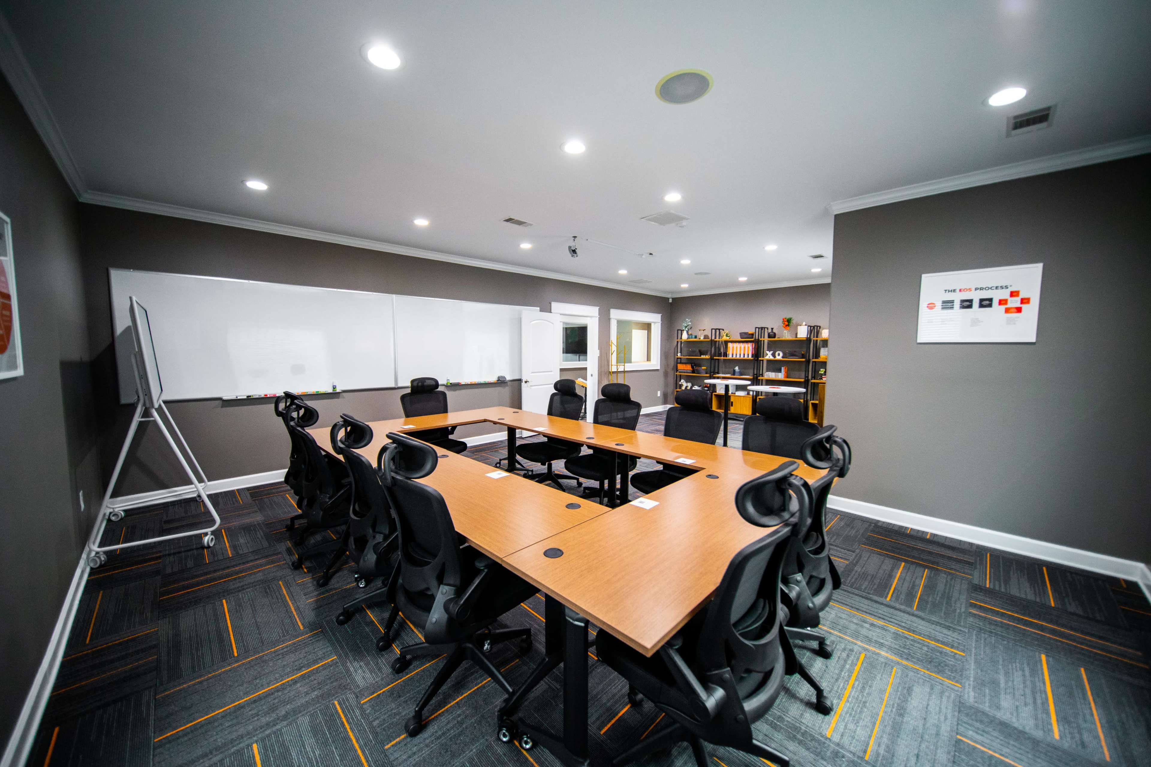 A modern conference room features a large wooden table surrounded by black chairs, with whiteboards and shelves in the background.