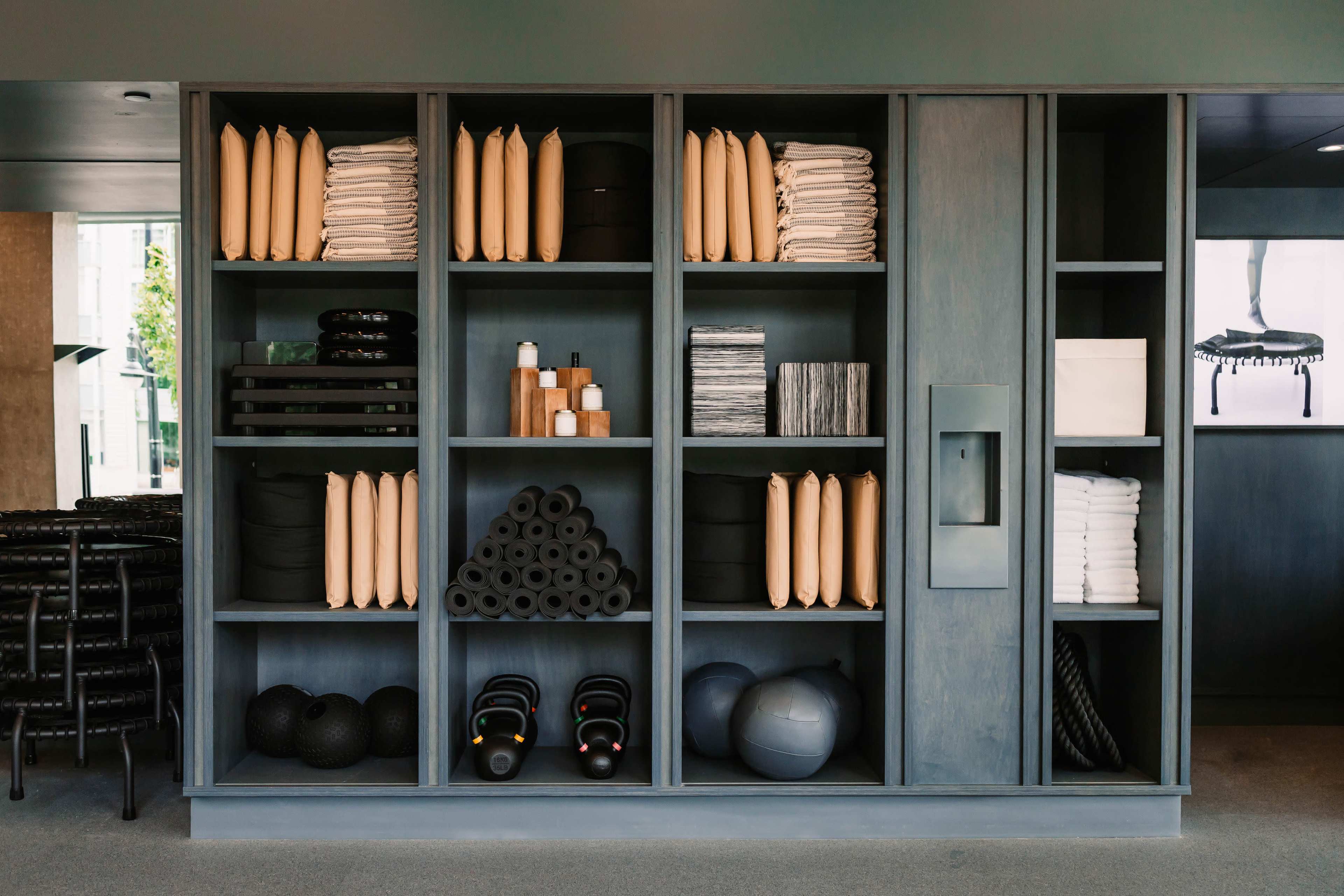 The image shows a neatly arranged storage unit filled with various fitness equipment and supplies, including rolled mats, weights, and towels.