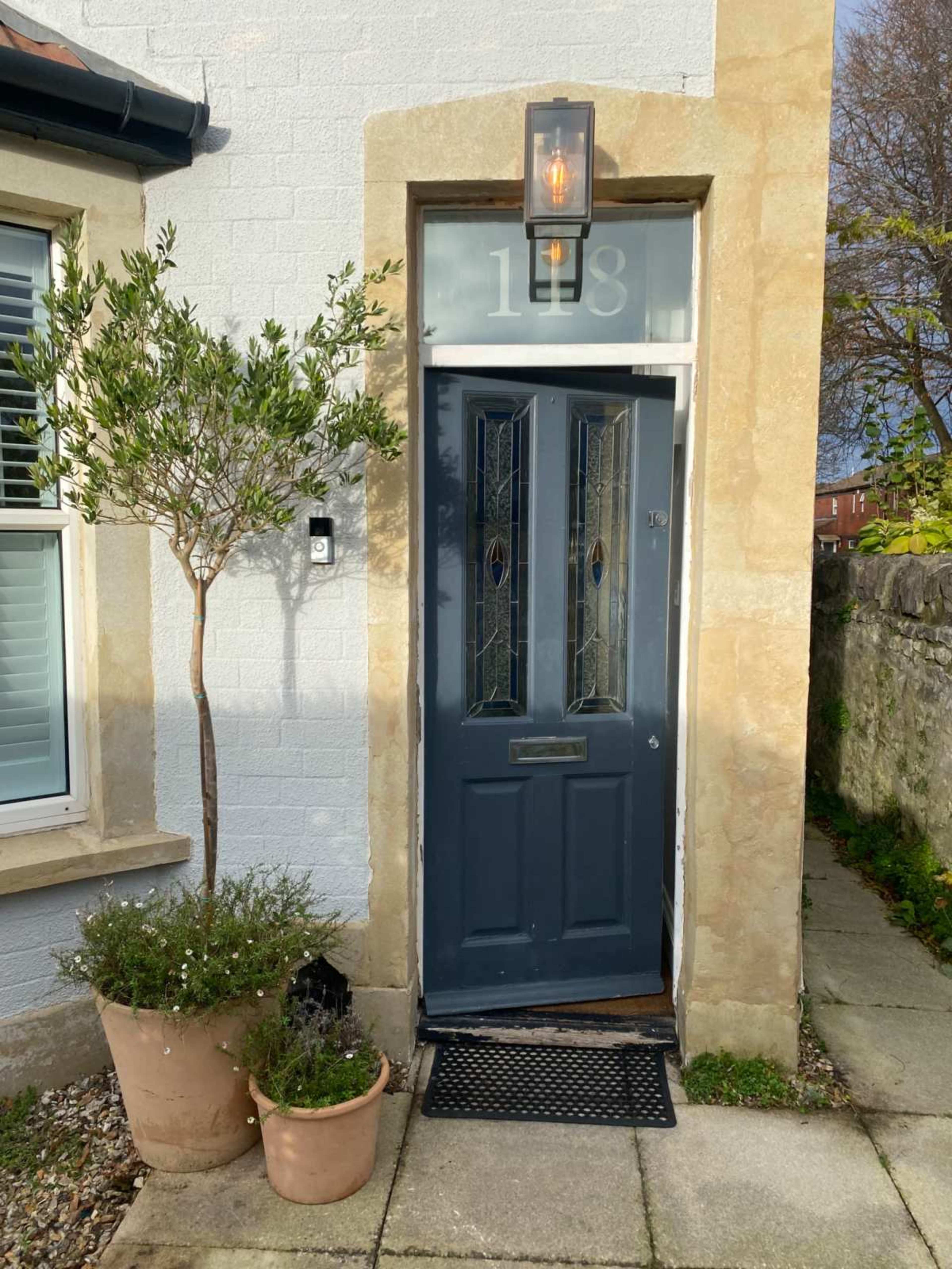 The image shows the entrance of a house with a blue front door, a light fixture above, and potted plants on either side of the doorway.
