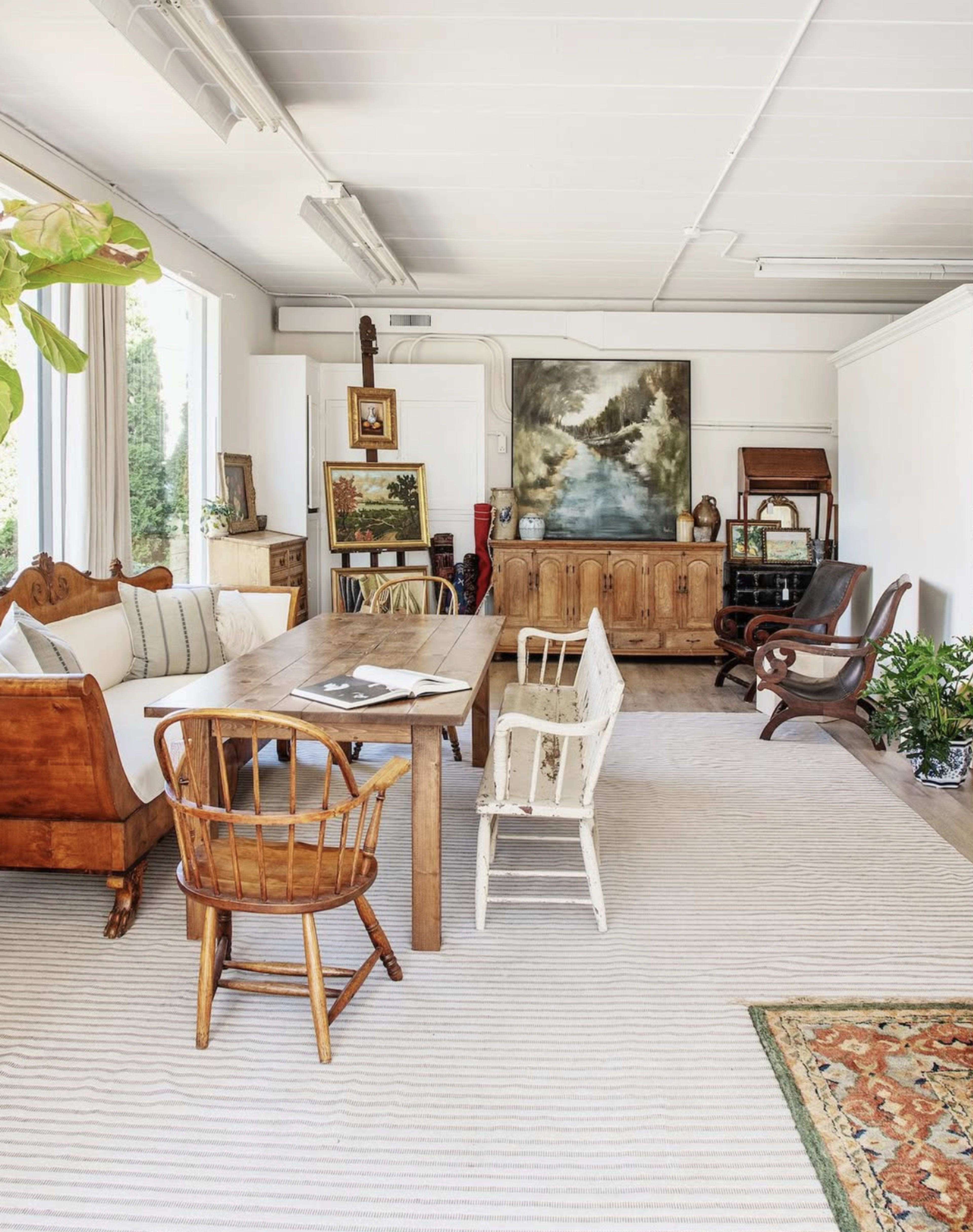A spacious room features a wooden dining table surrounded by vintage chairs, with a large painting and various decorative items on the walls.