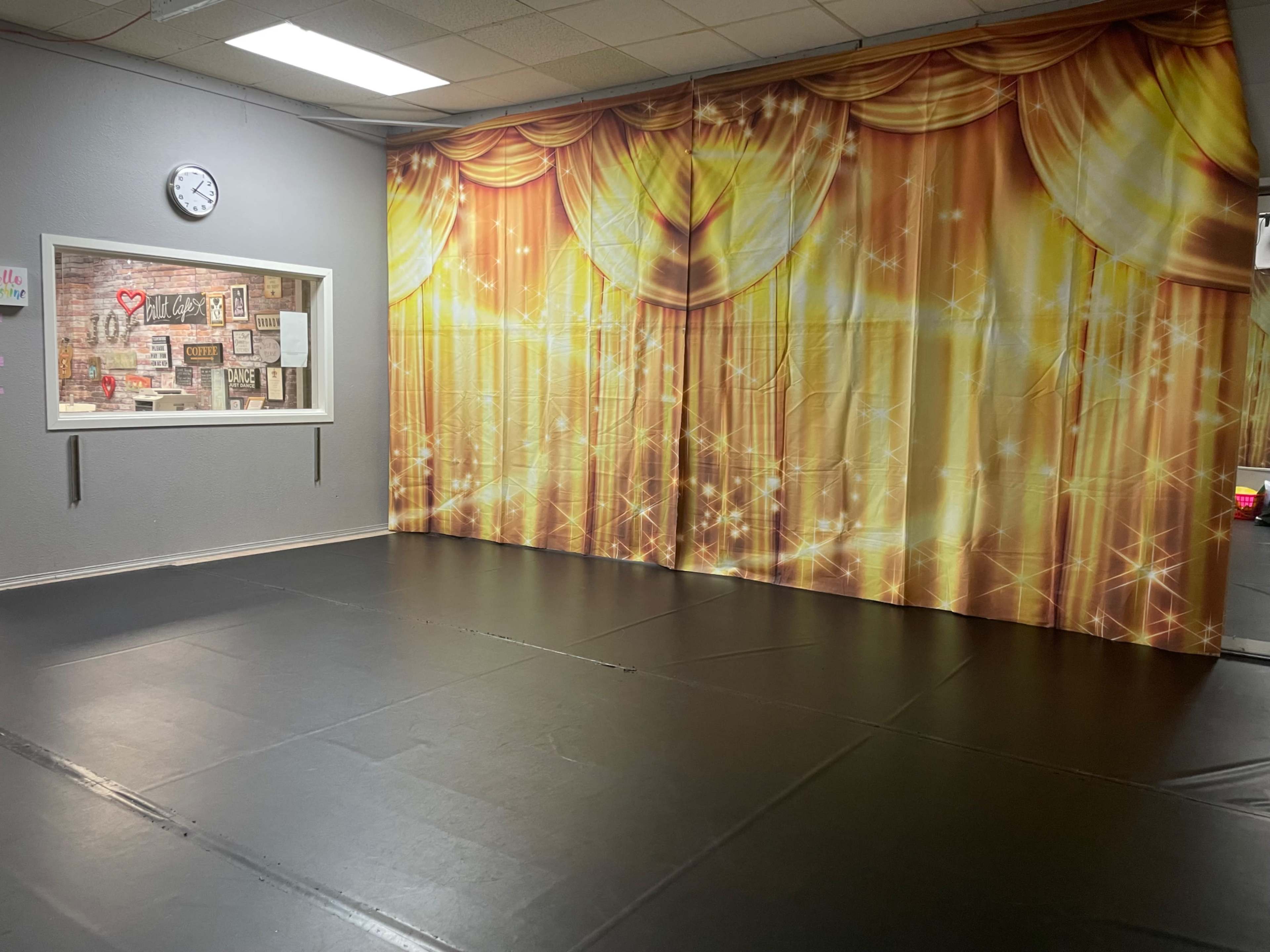 A dance studio with a black floor and a golden curtain backdrop, along with a wall-mounted clock and a window displaying various posters.