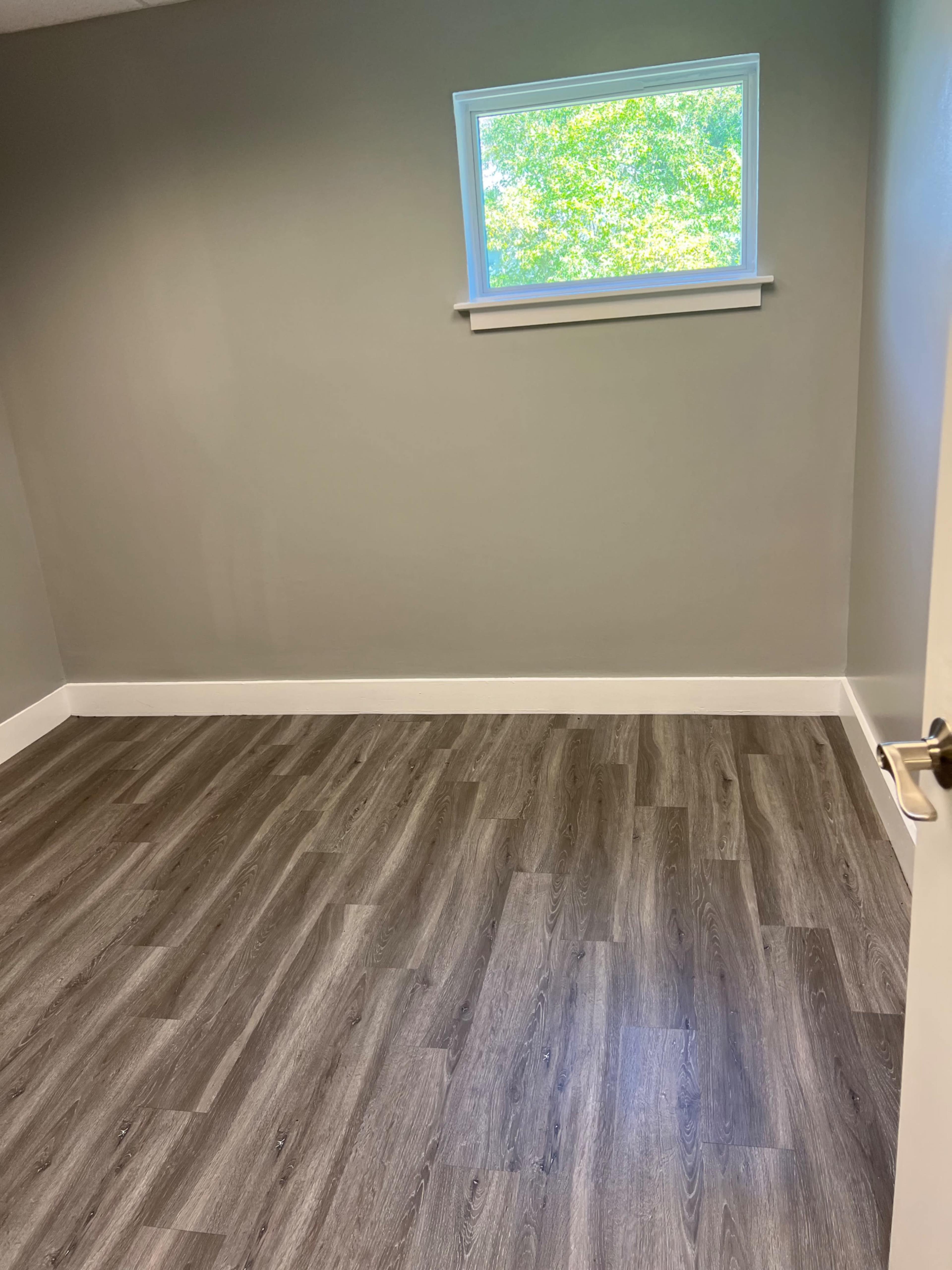 An empty room with gray walls, a window allowing natural light to enter, and dark wood-like flooring.