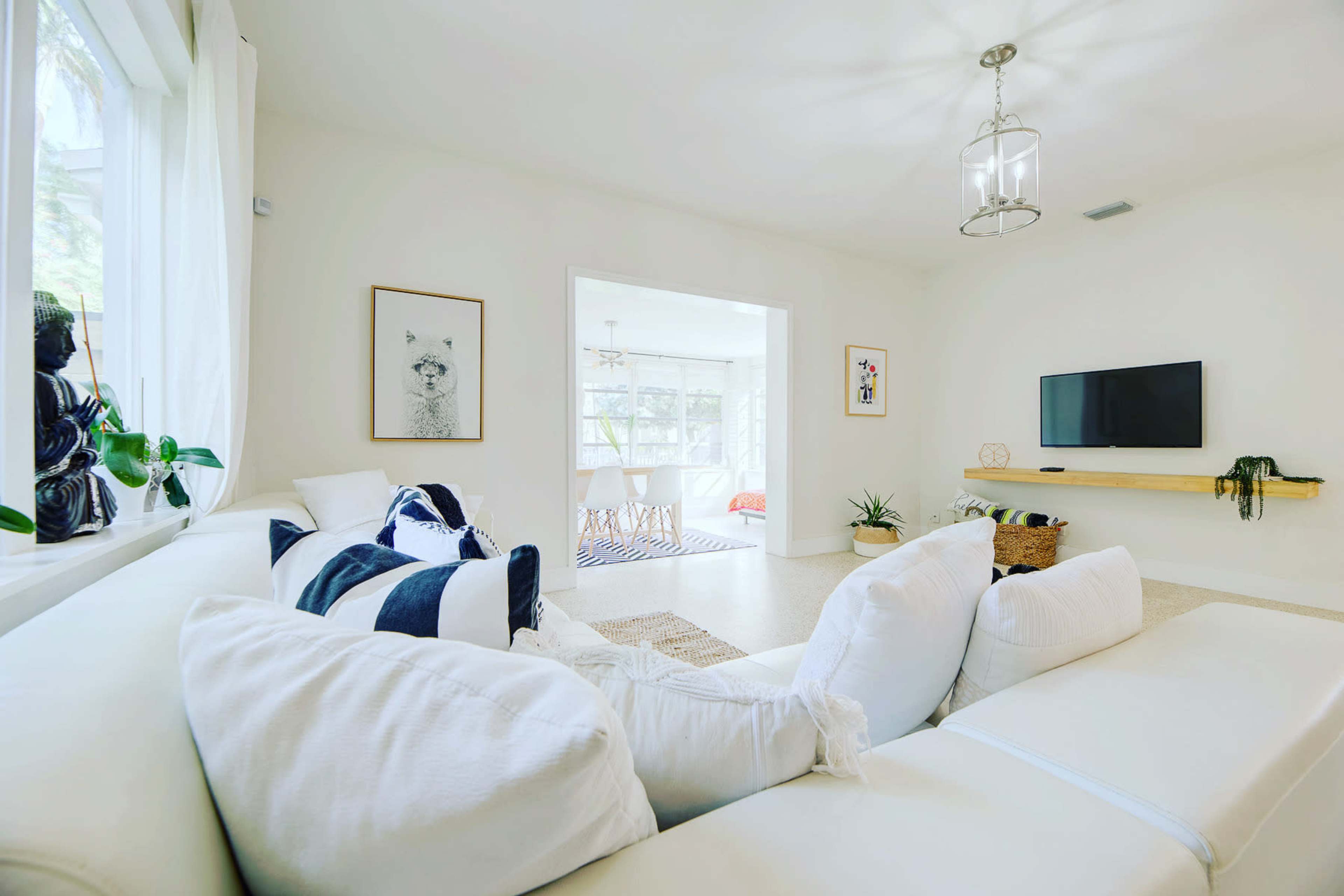 The image shows a bright, modern living room with a white sectional couch, a television mounted on the wall, and a view into a second room with large windows.