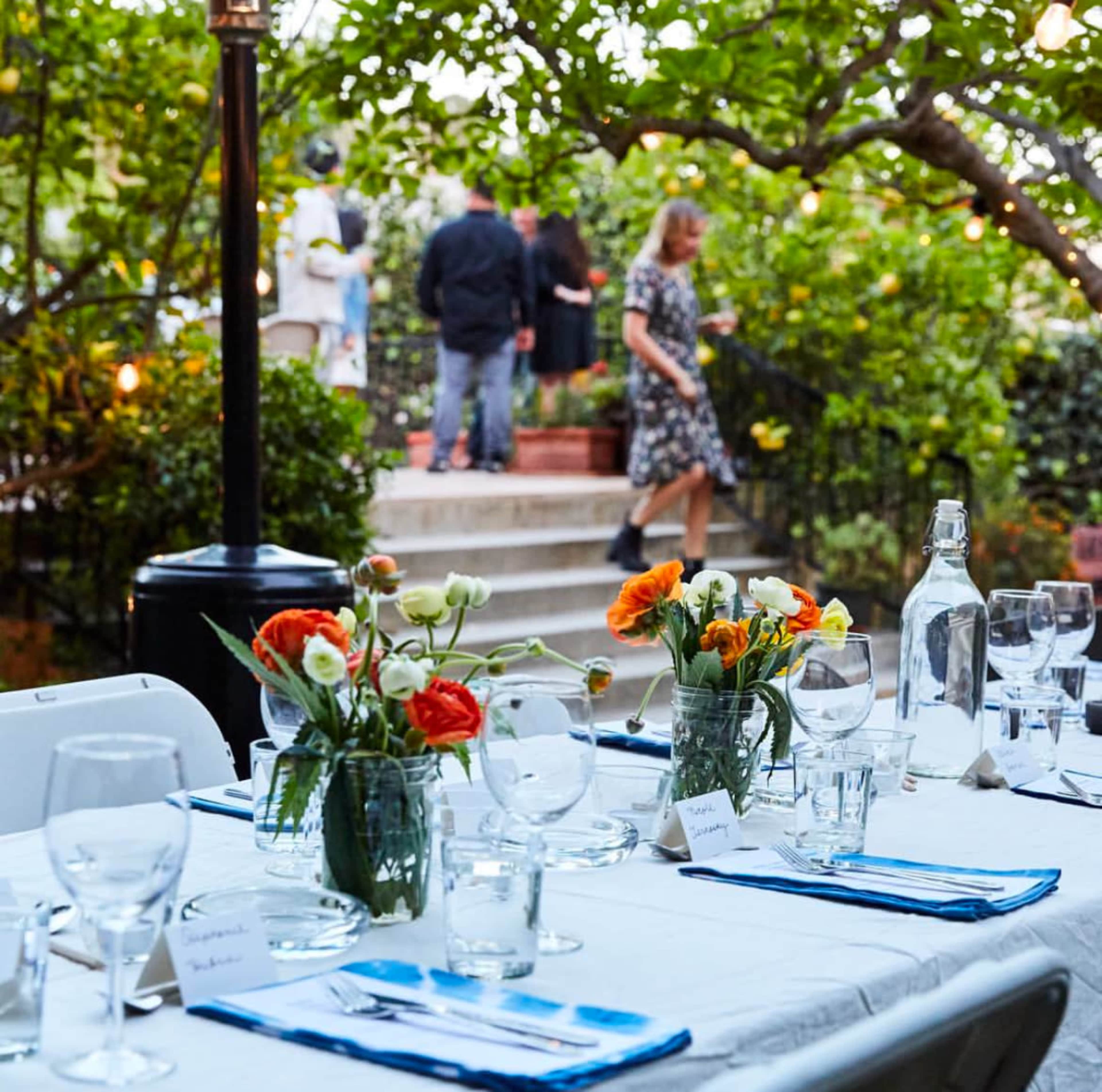 A beautifully set outdoor dining table with flowers in jars, surrounded by people mingling in a garden.