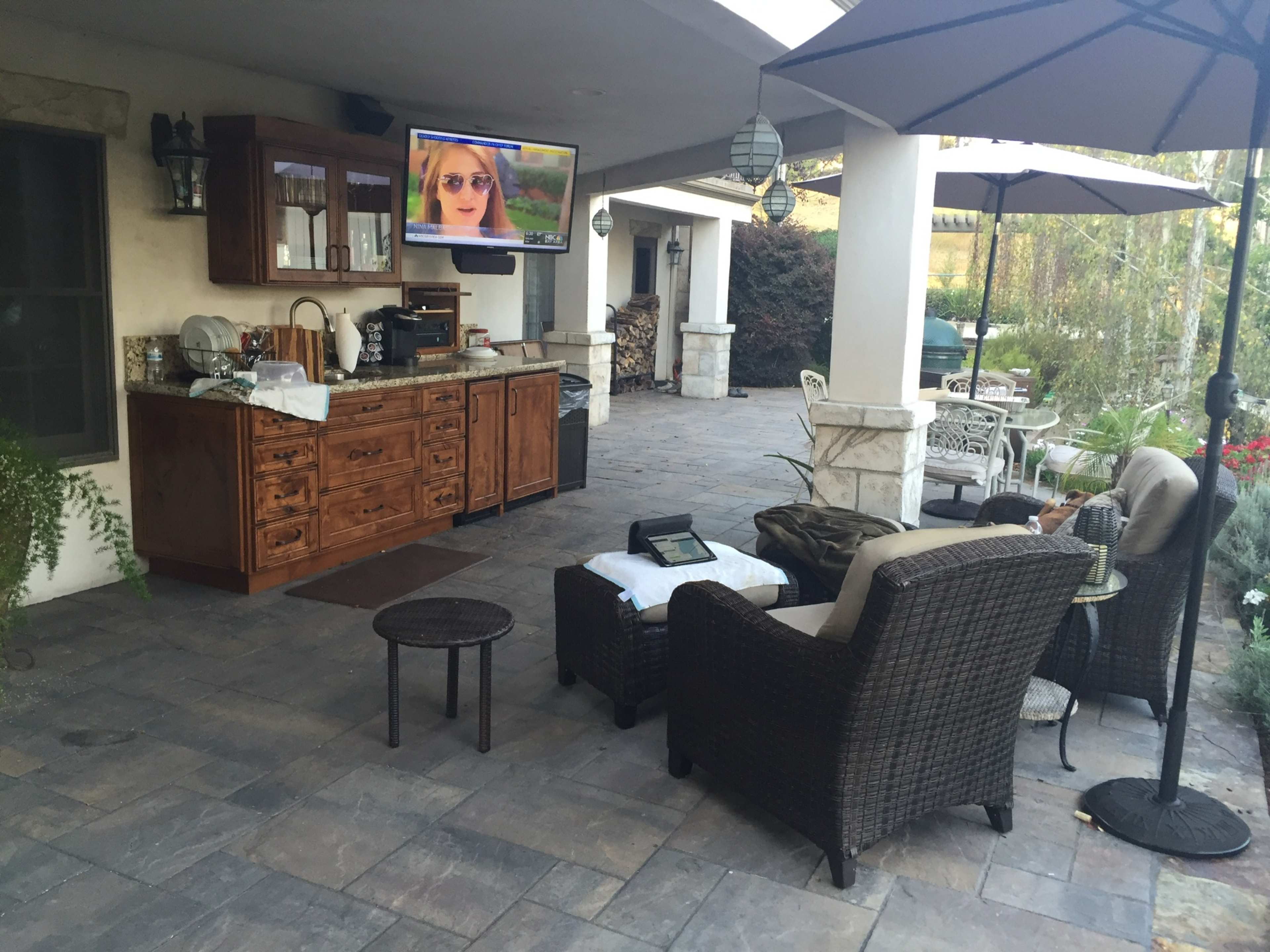 A covered outdoor patio with a stone floor, a wooden cabinet, a television mounted on the wall, and various seating arrangements under umbrellas.