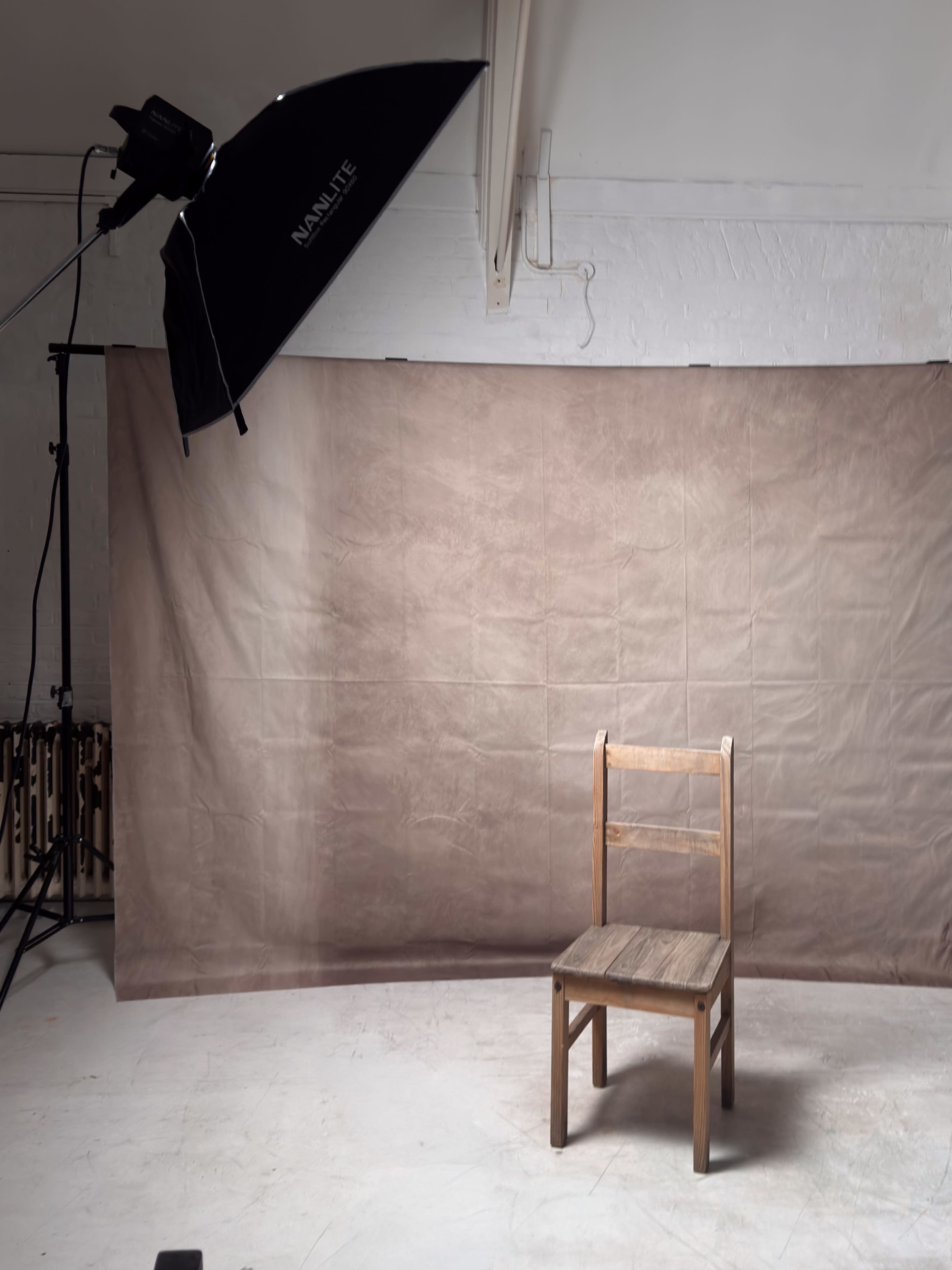 The image depicts a simple wooden chair positioned in front of a plain brown backdrop, with studio lighting set up nearby.